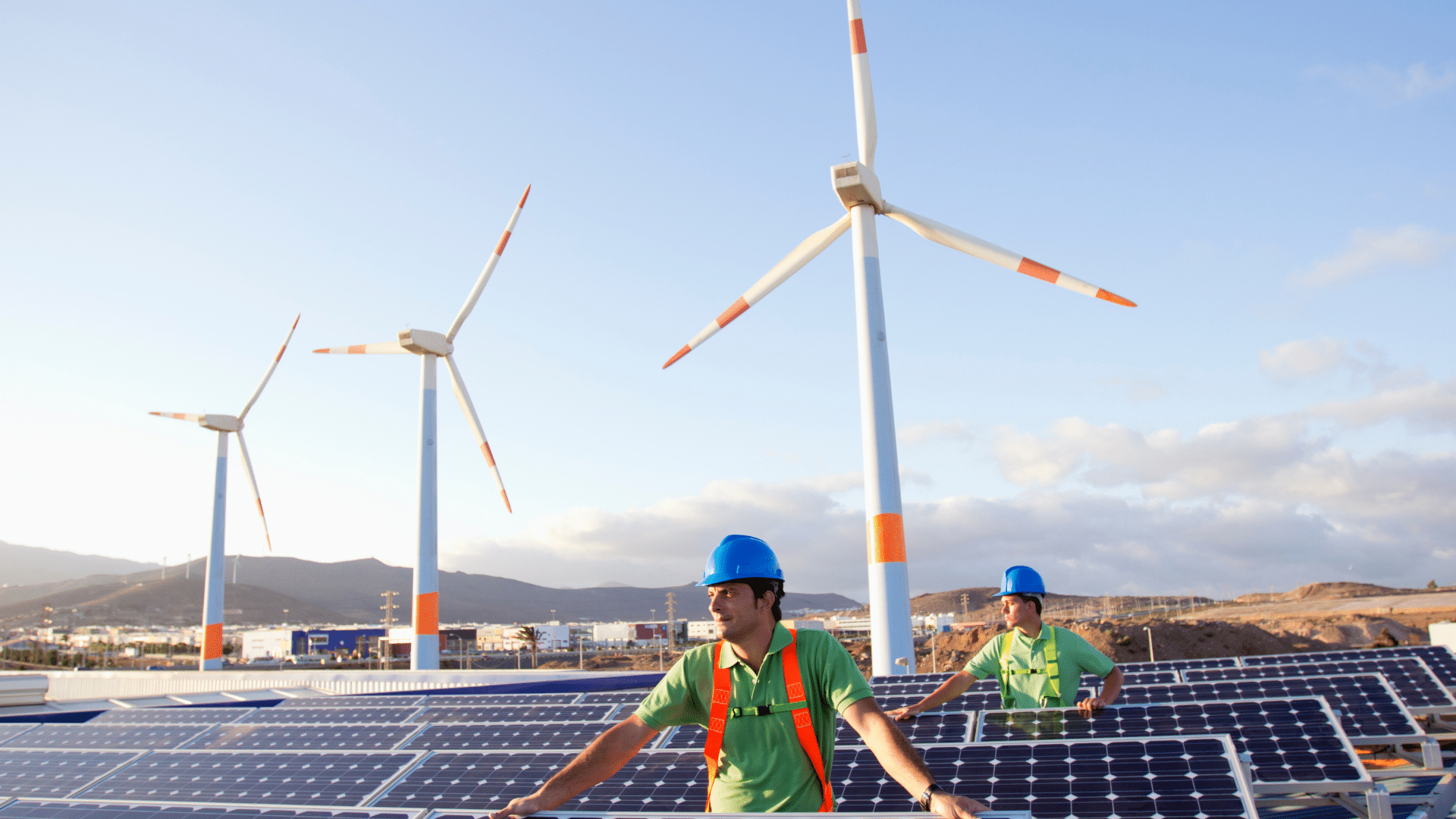 Website blog covers – Energy (40) Two workers in helmets stand by solar panels with wind turbines in the background on a clear day.