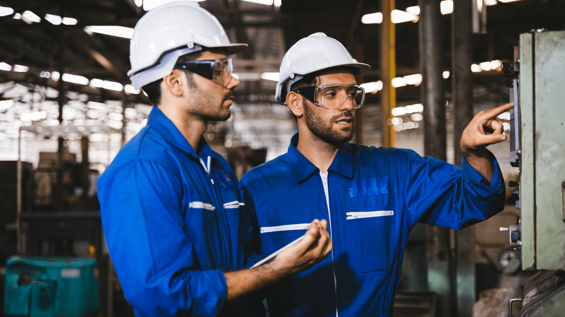 Two workers in blue uniforms and hard hats operate machinery in a factory.