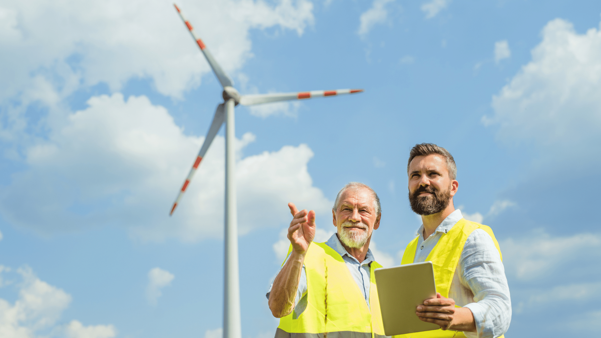 Two men in safety vests stand near a wind turbine under a blue sky with clouds.
