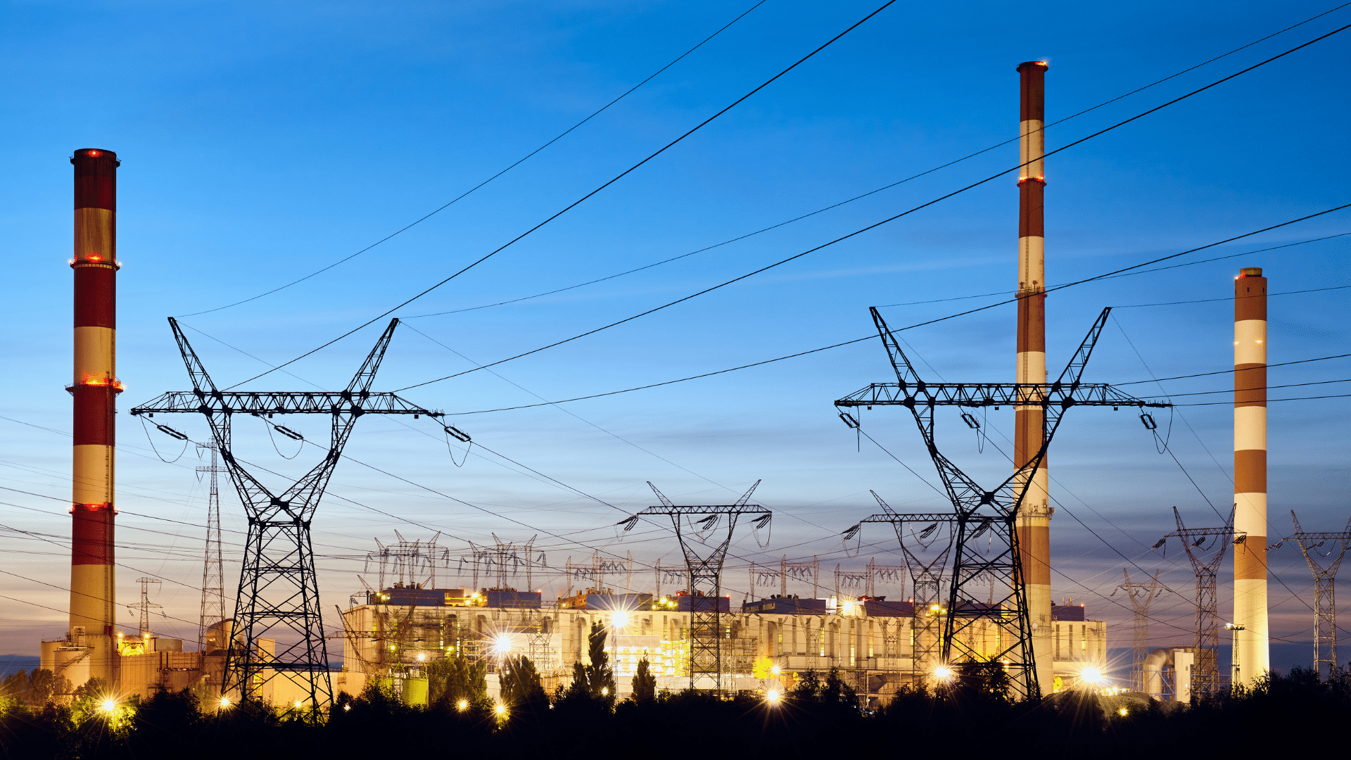 Power plant with smokestacks and electricity pylons at dusk.