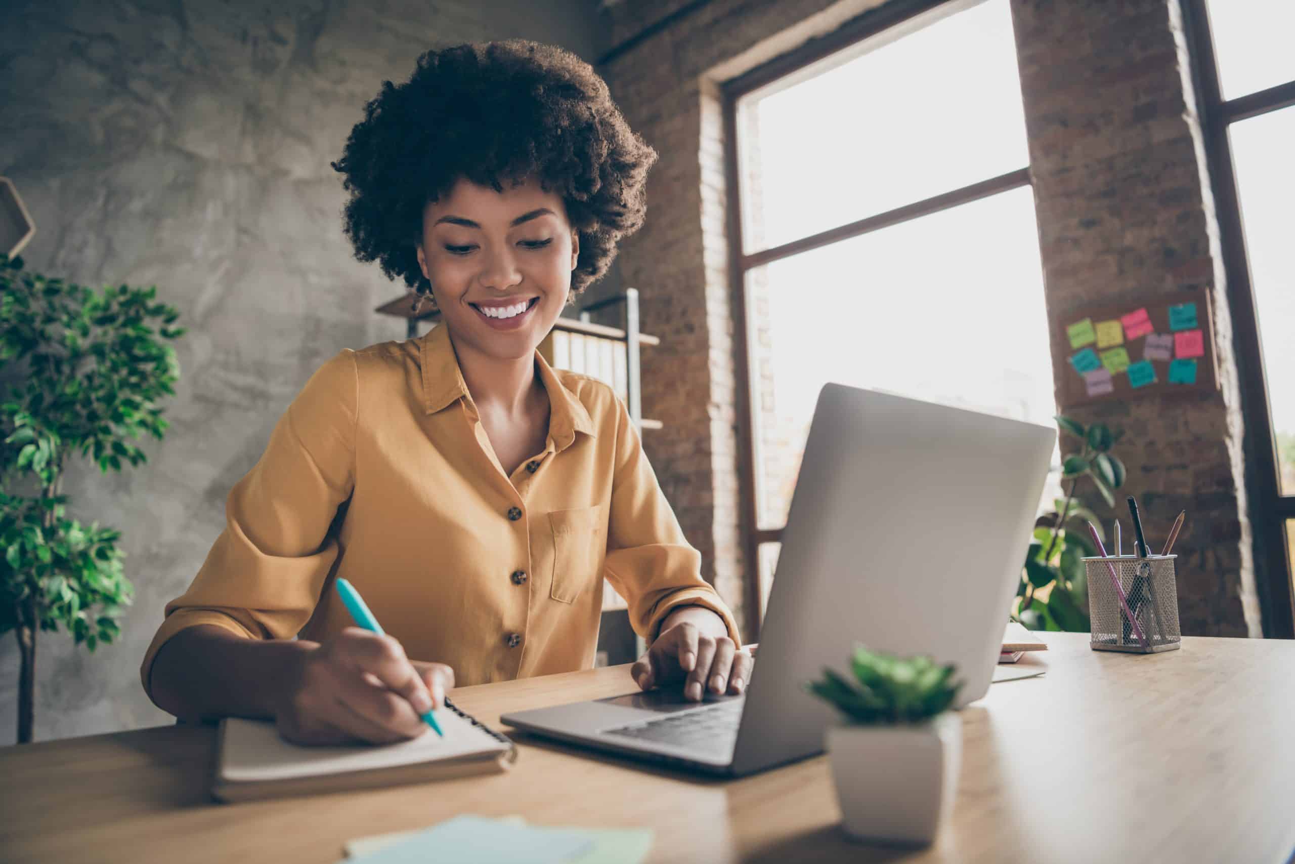 A woman sat at a desk in front of a laptop writing in a notebook