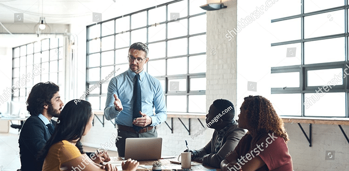 A man leads a discussion with four people seated around a table in a modern office setting.
