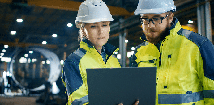 Two workers in safety gear look at a laptop in an industrial setting.