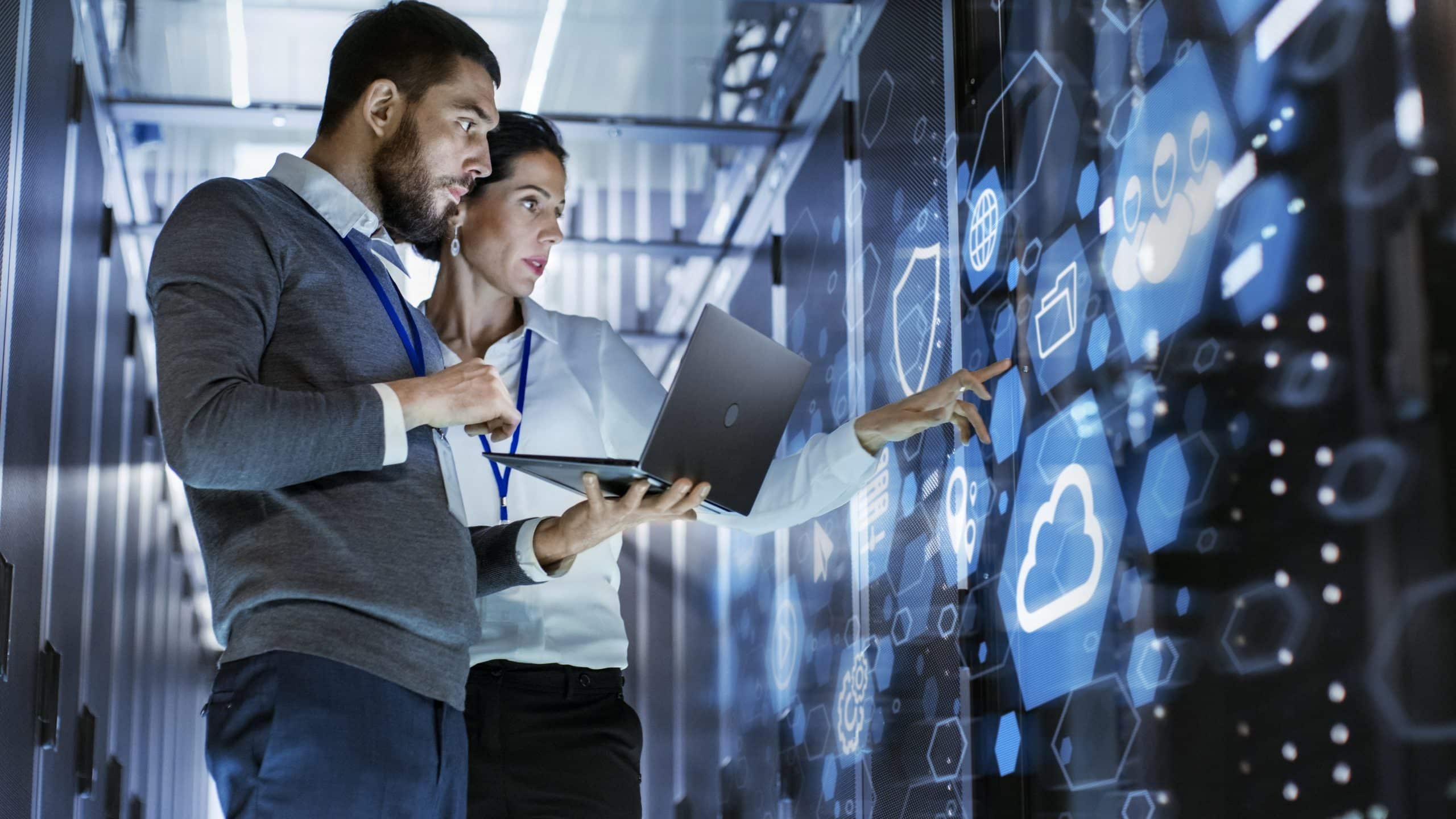 Two people using a laptop and interacting with a digital touchscreen in a server room.