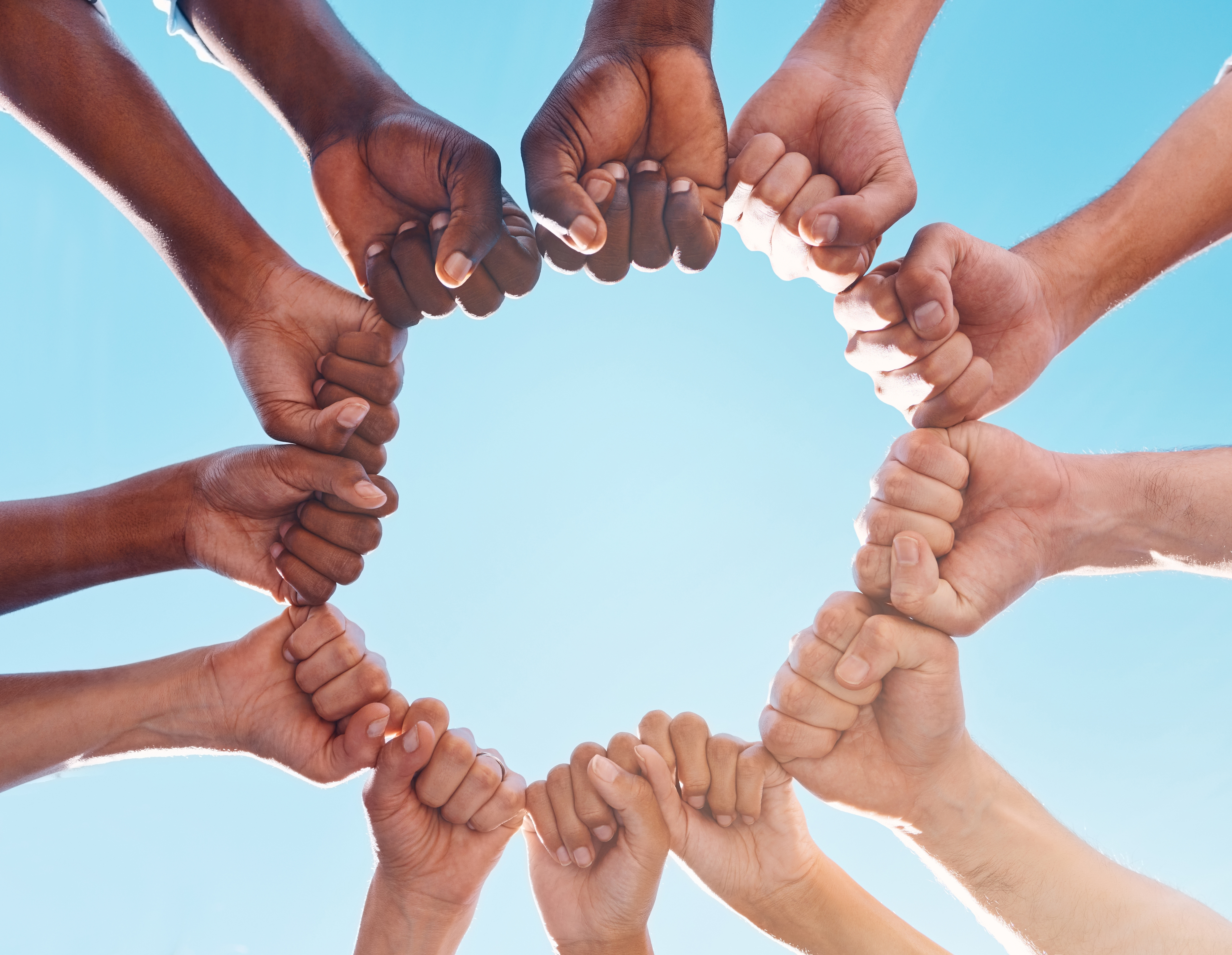 A circle of diverse hands with fists touching against a blue sky.