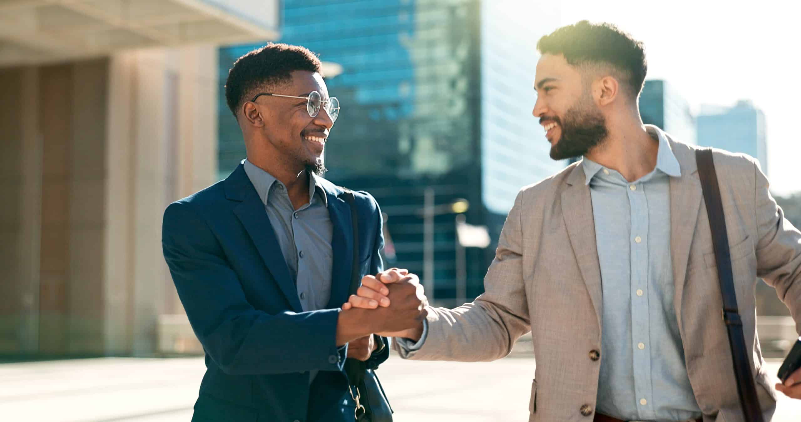 Two businessmen smiling and shaking hands outdoors in a city setting.