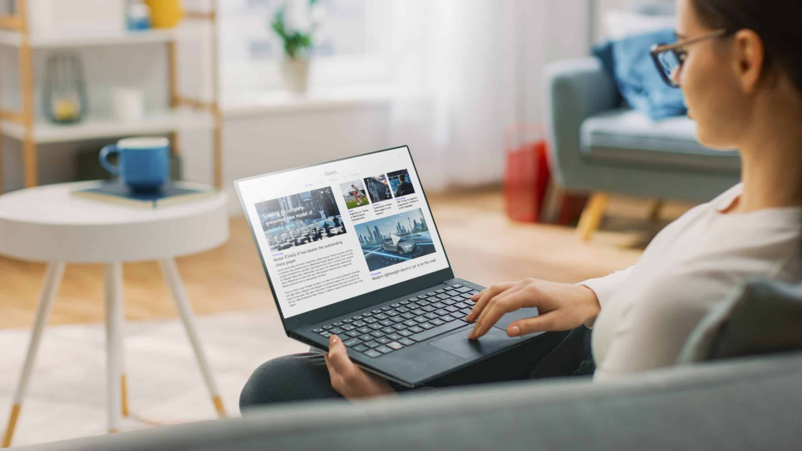A woman sits on a couch reading blogs on a laptop in a cozy living room.