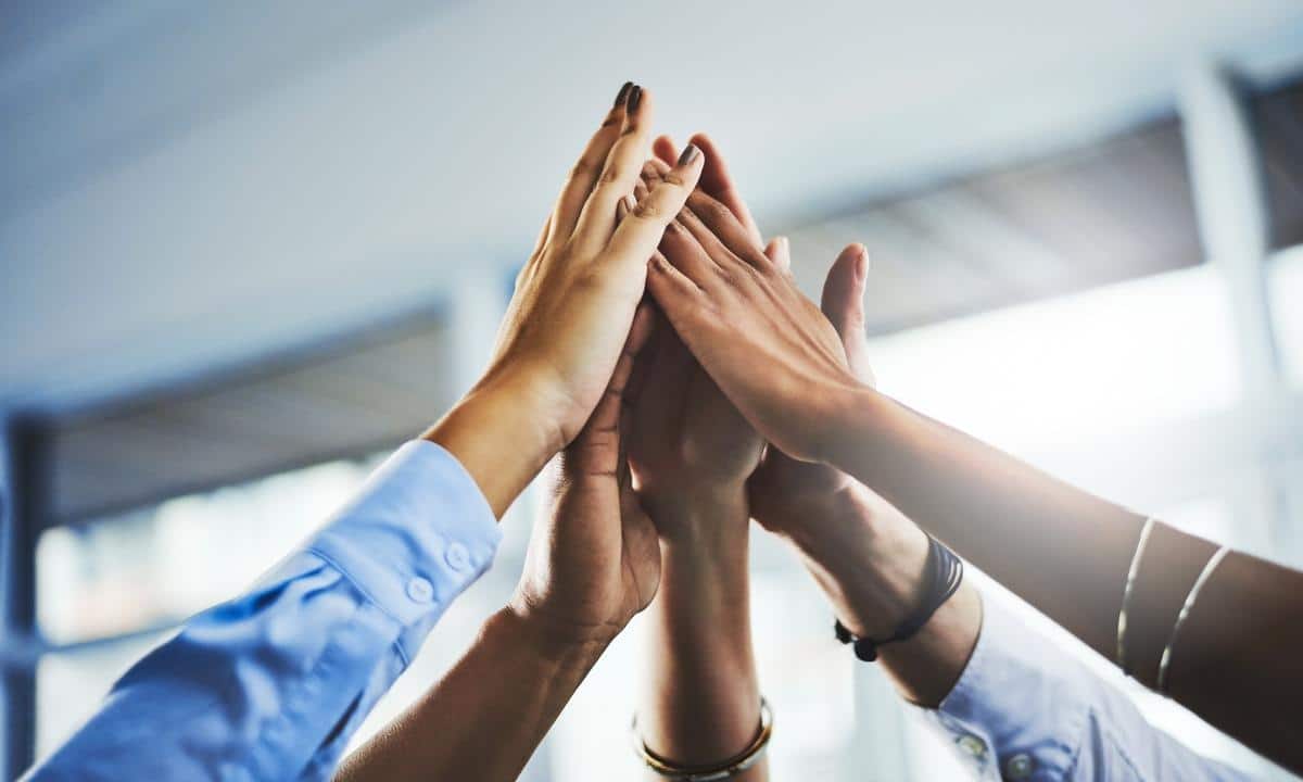 Five people in business attire join hands for a group high five in an office setting.