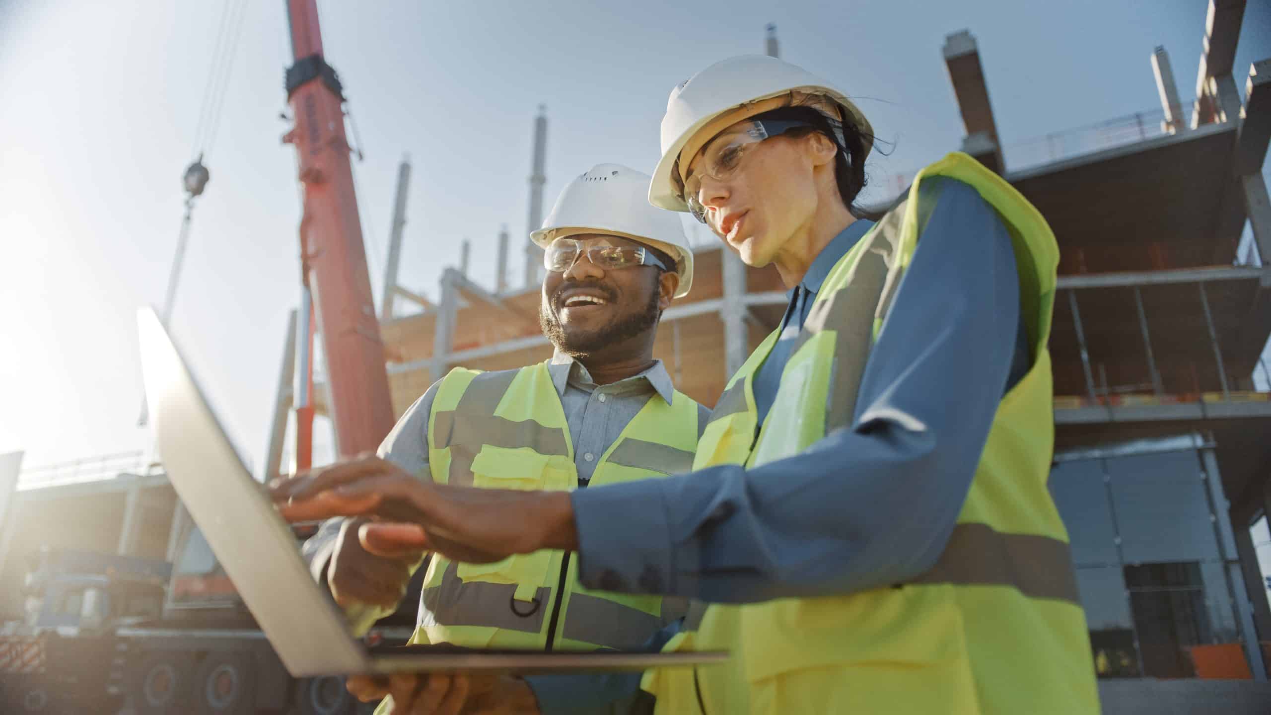 Two construction workers in safety gear look at a laptop on a building site.