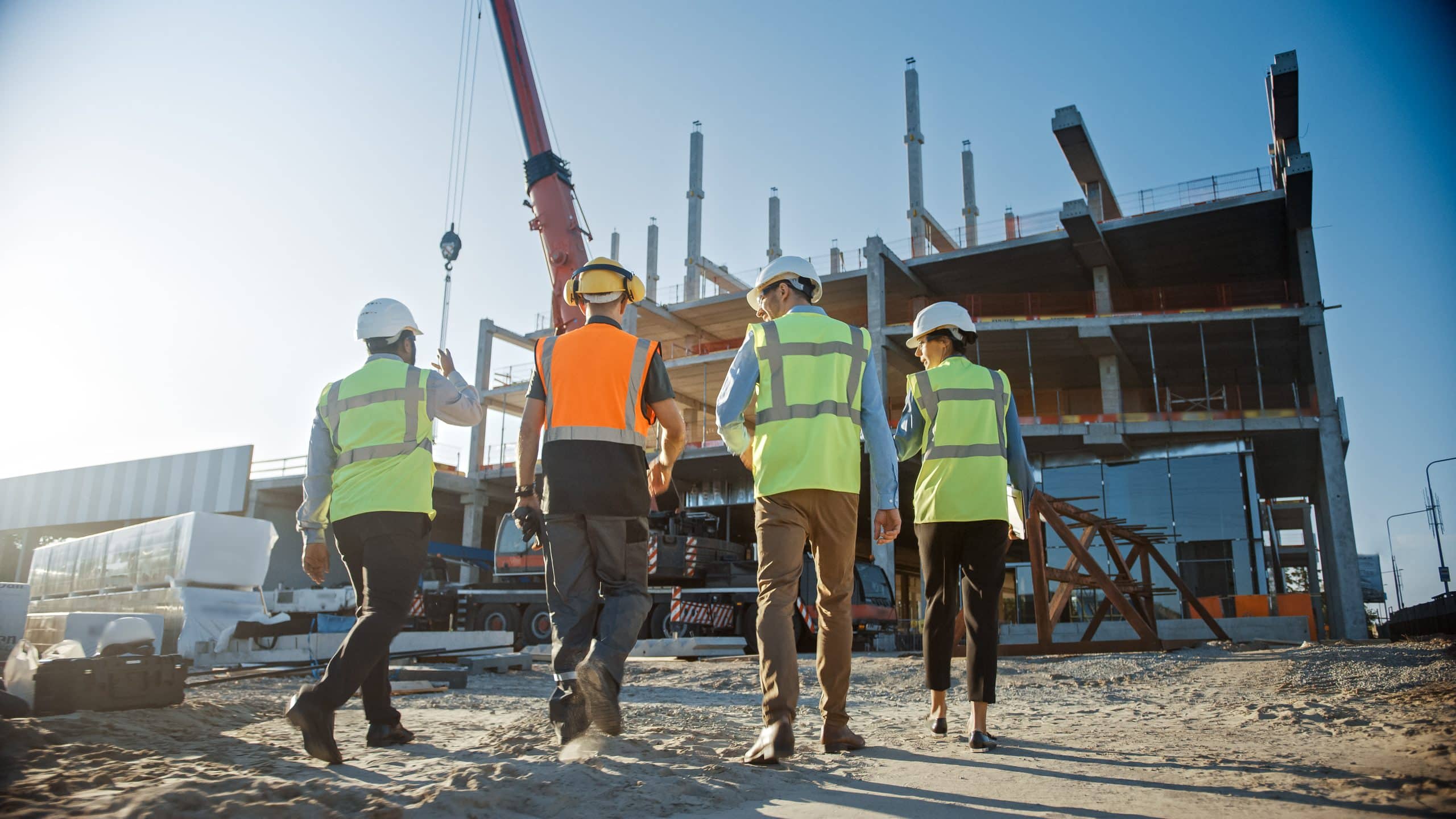 Four construction workers in safety vests and helmets walk toward a building under construction.