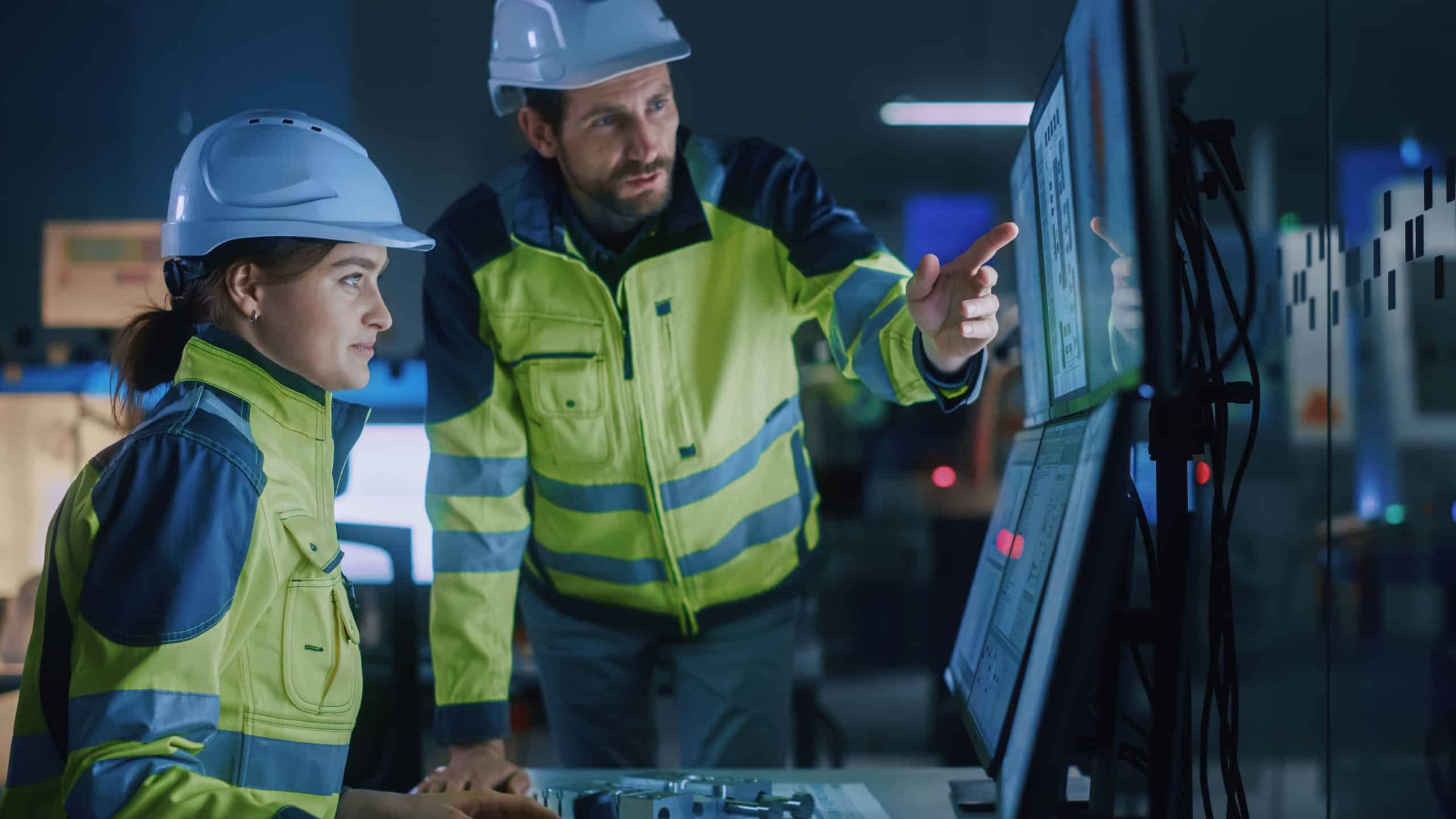 Two construction workers in safety gear look at computer screens in an office.