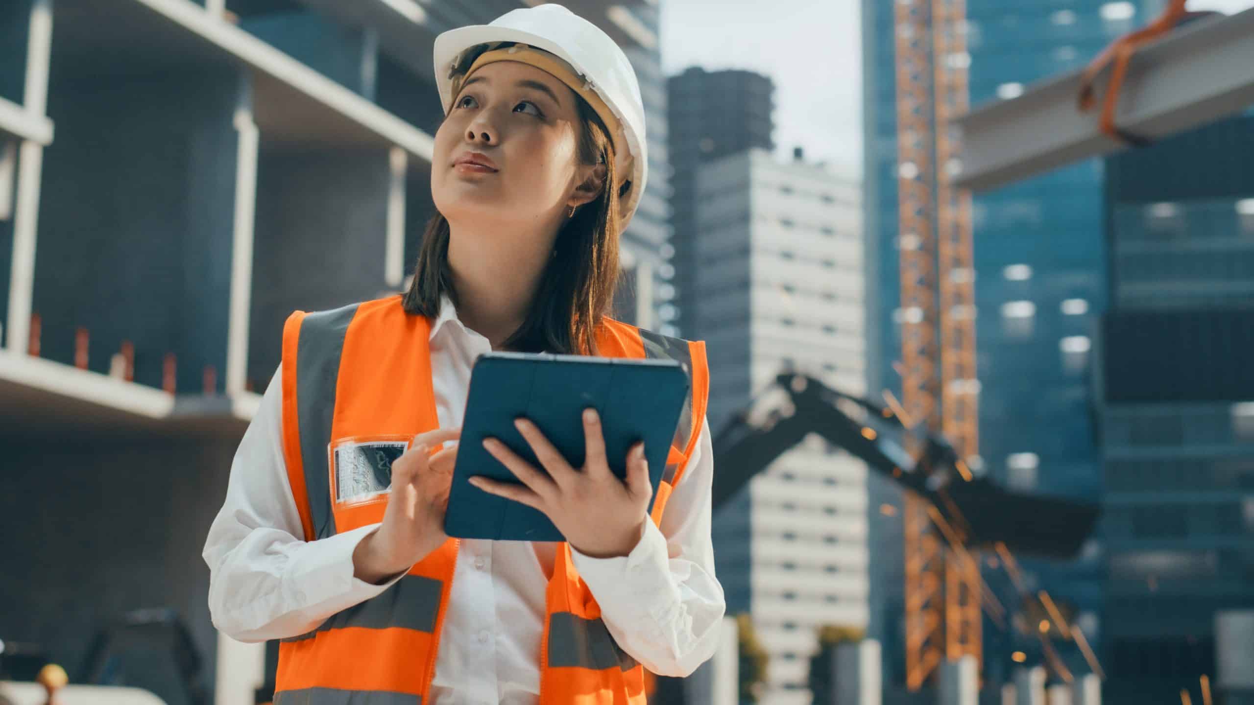 Woman in a safety vest and hard hat using a tablet at a construction site.