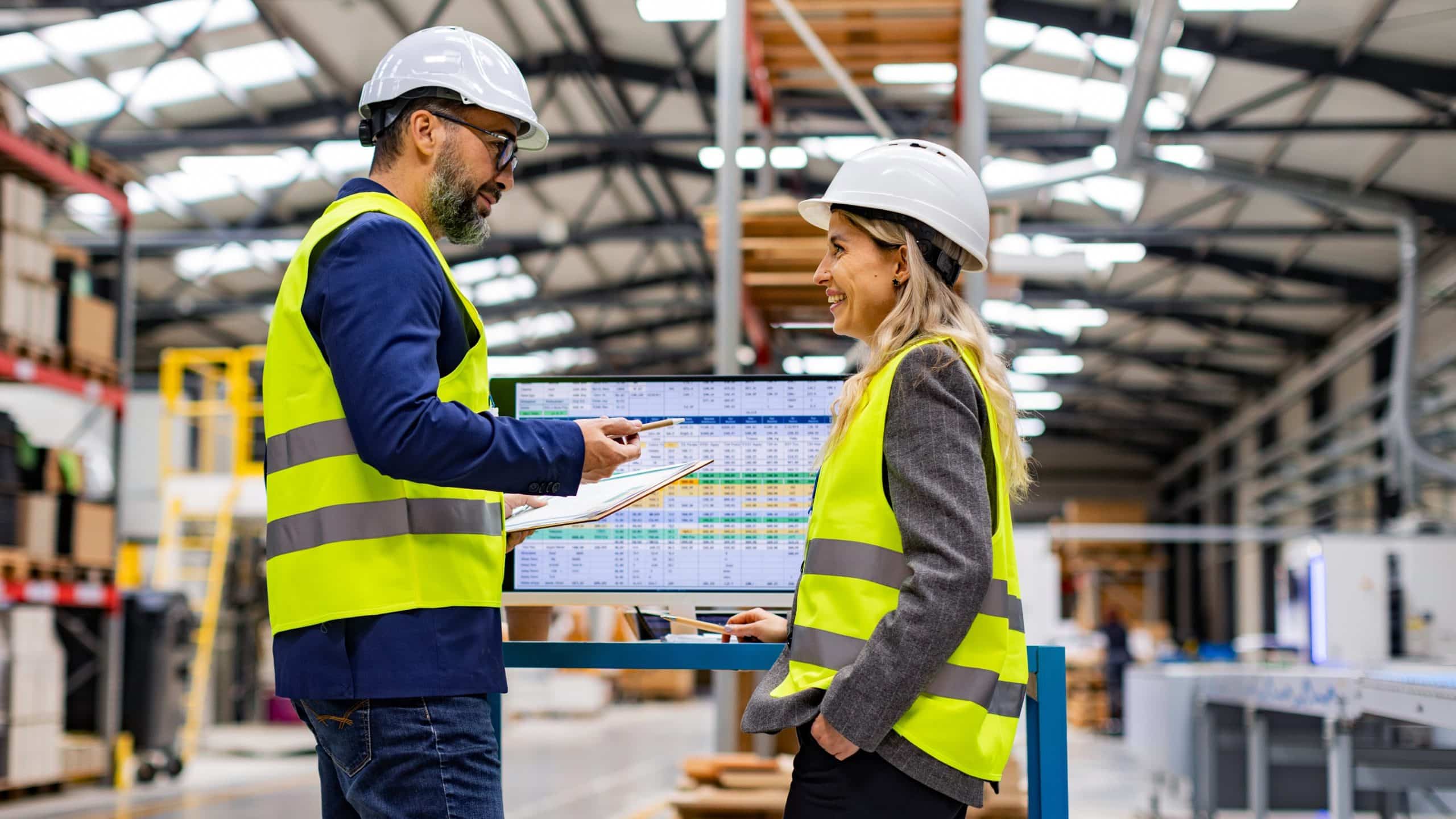 Two workers in safety vests and helmets talk in a warehouse with a computer screen behind them.