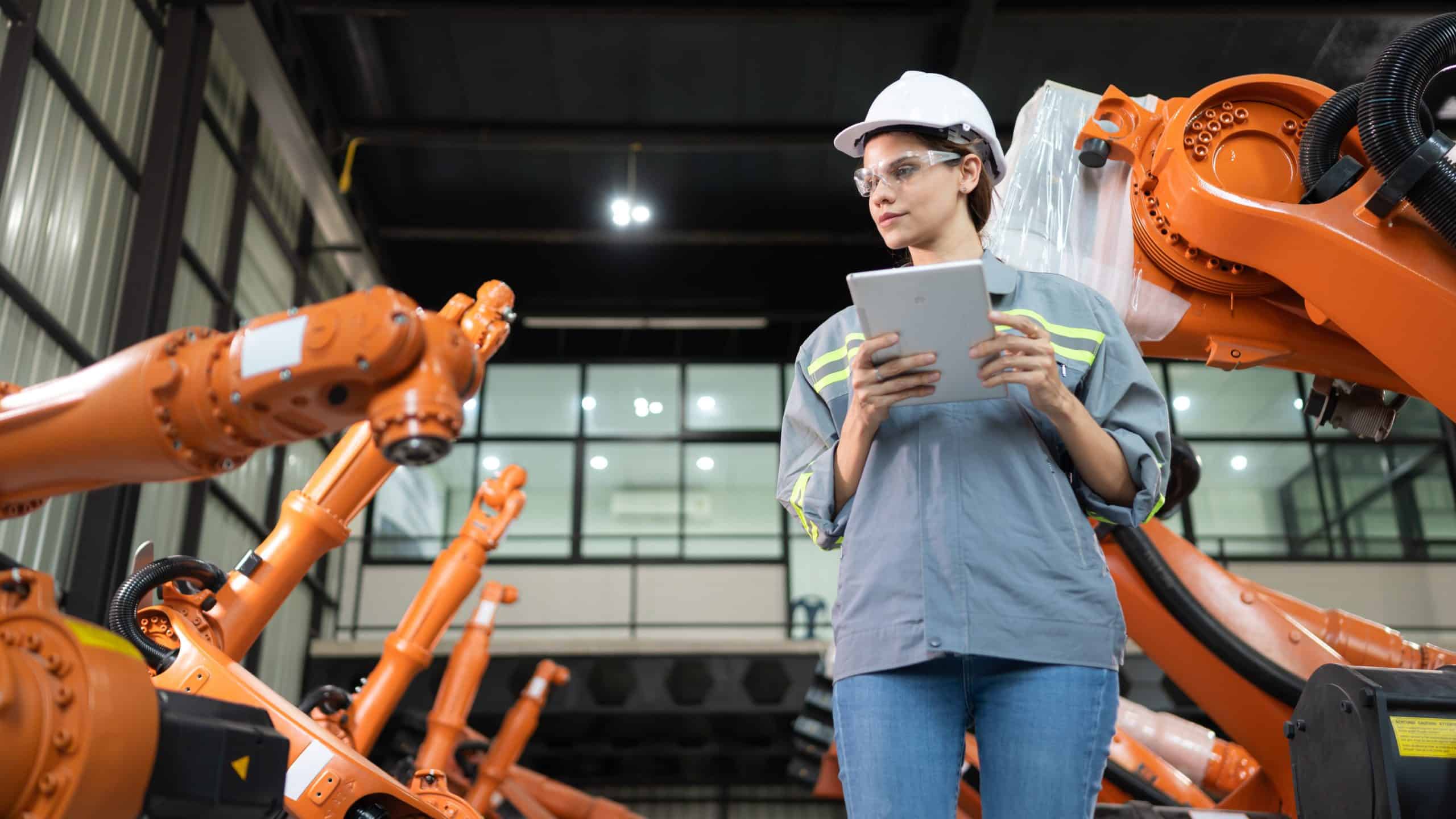 Woman in a hard hat uses a tablet to inspect industrial robotic arms in a factory setting.