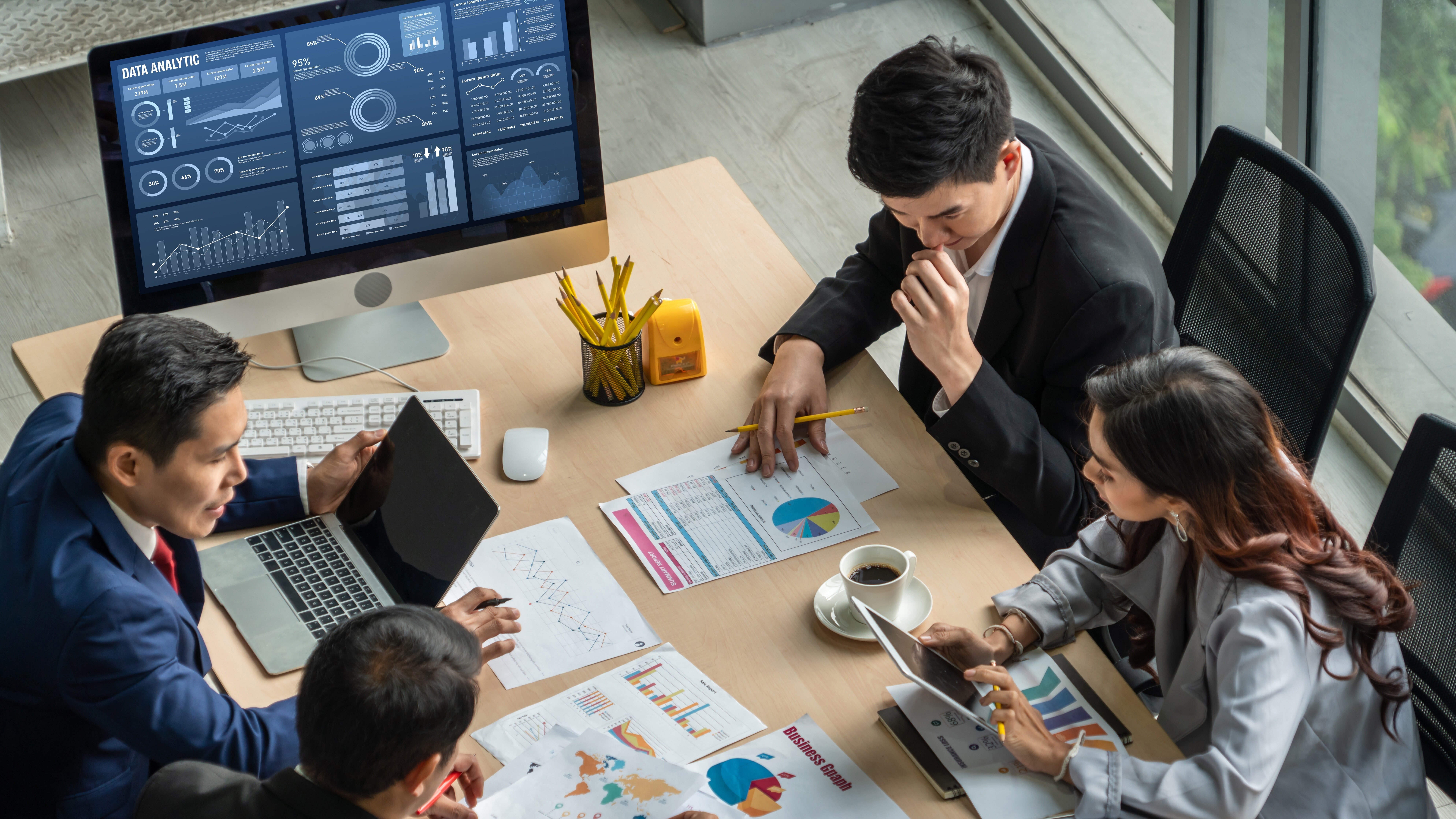 Four people in a meeting room discussing charts and graphs with a data analytics screen on a monitor.