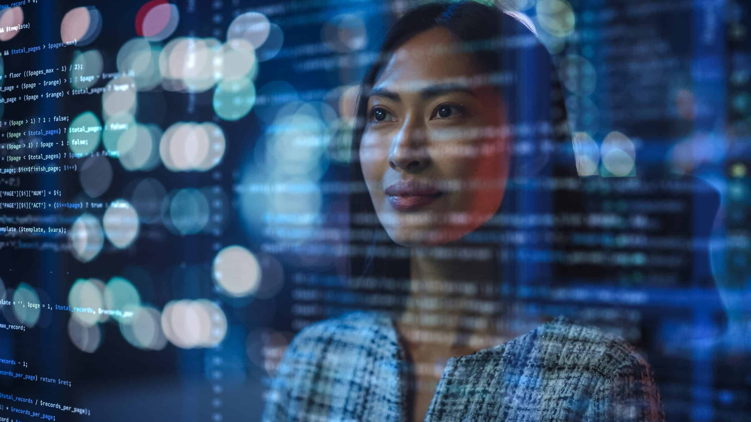 A woman looks at computer code on a screen, with city lights in the background.