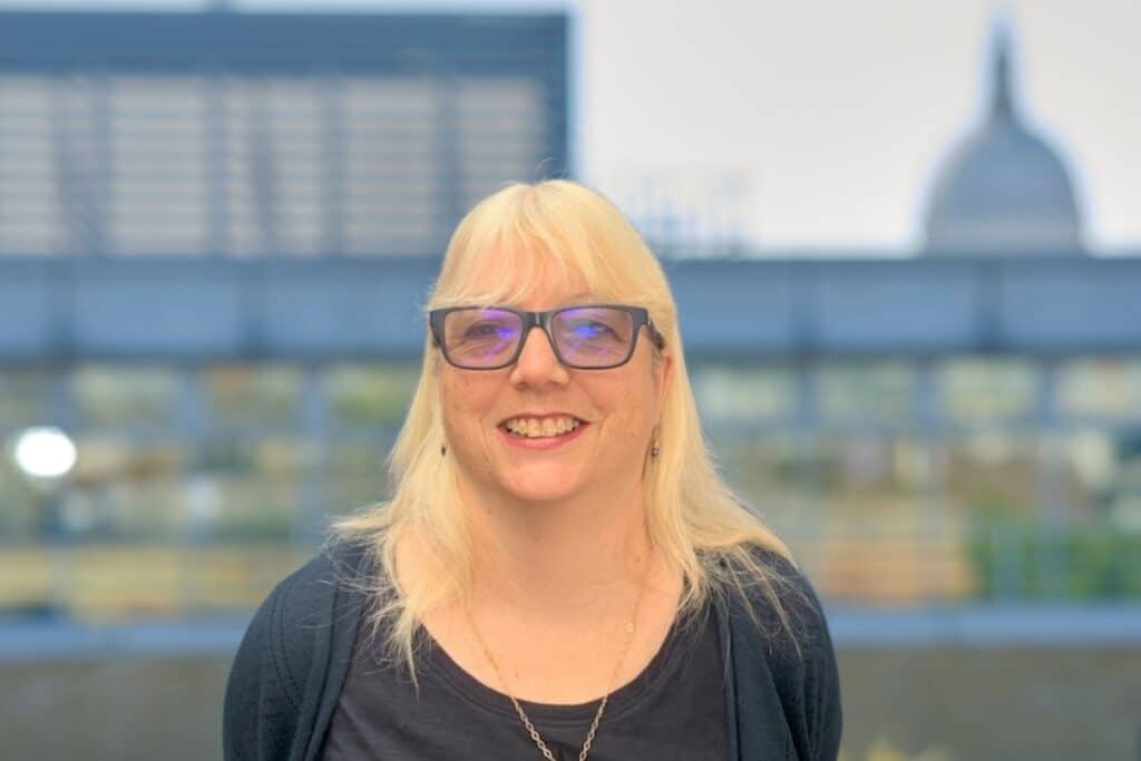 Smiling woman with glasses and blonde hair, outdoors with a cityscape background.