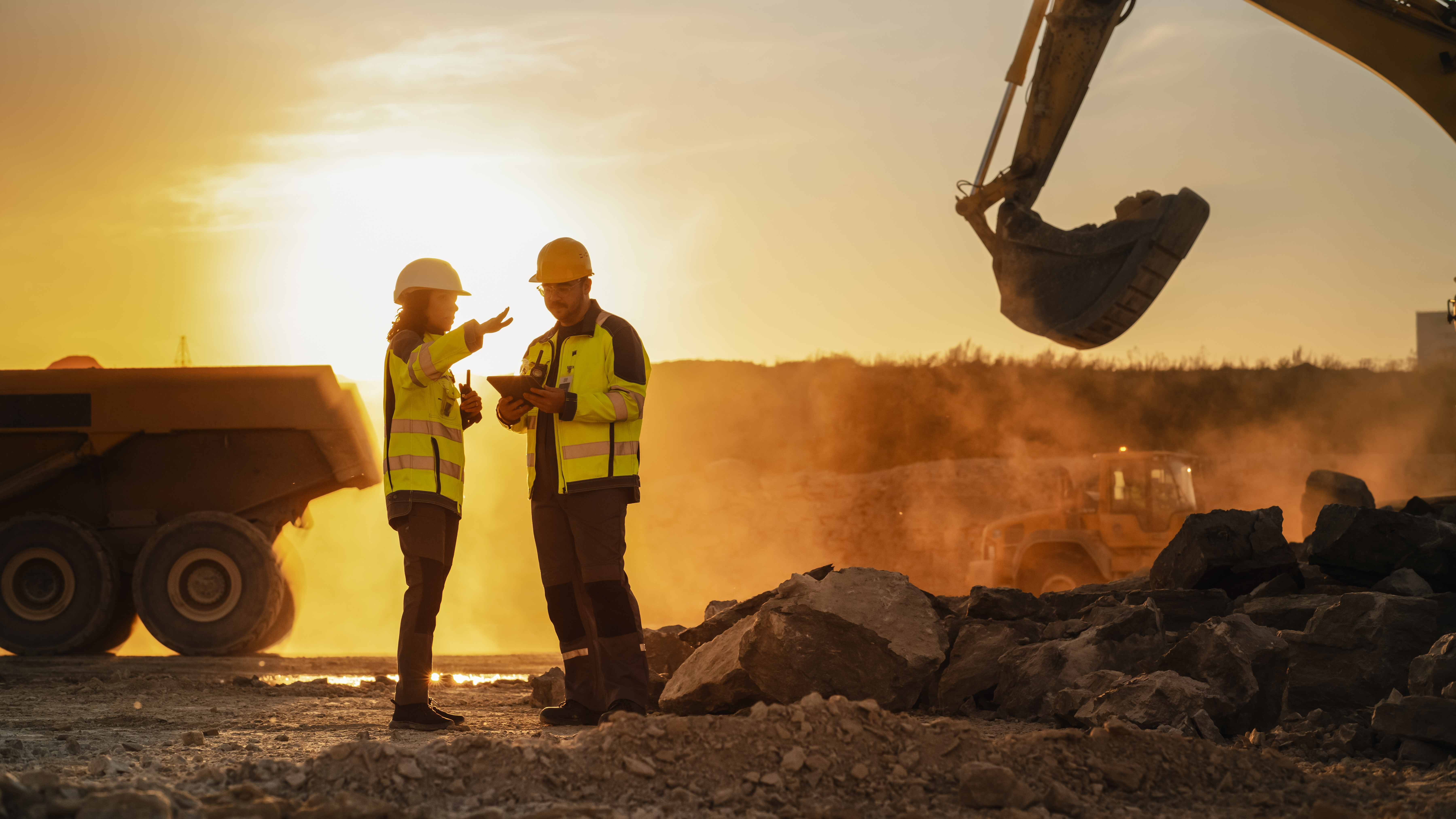 Two workers in safety gear stand at a mining site with heavy machinery at sunset.