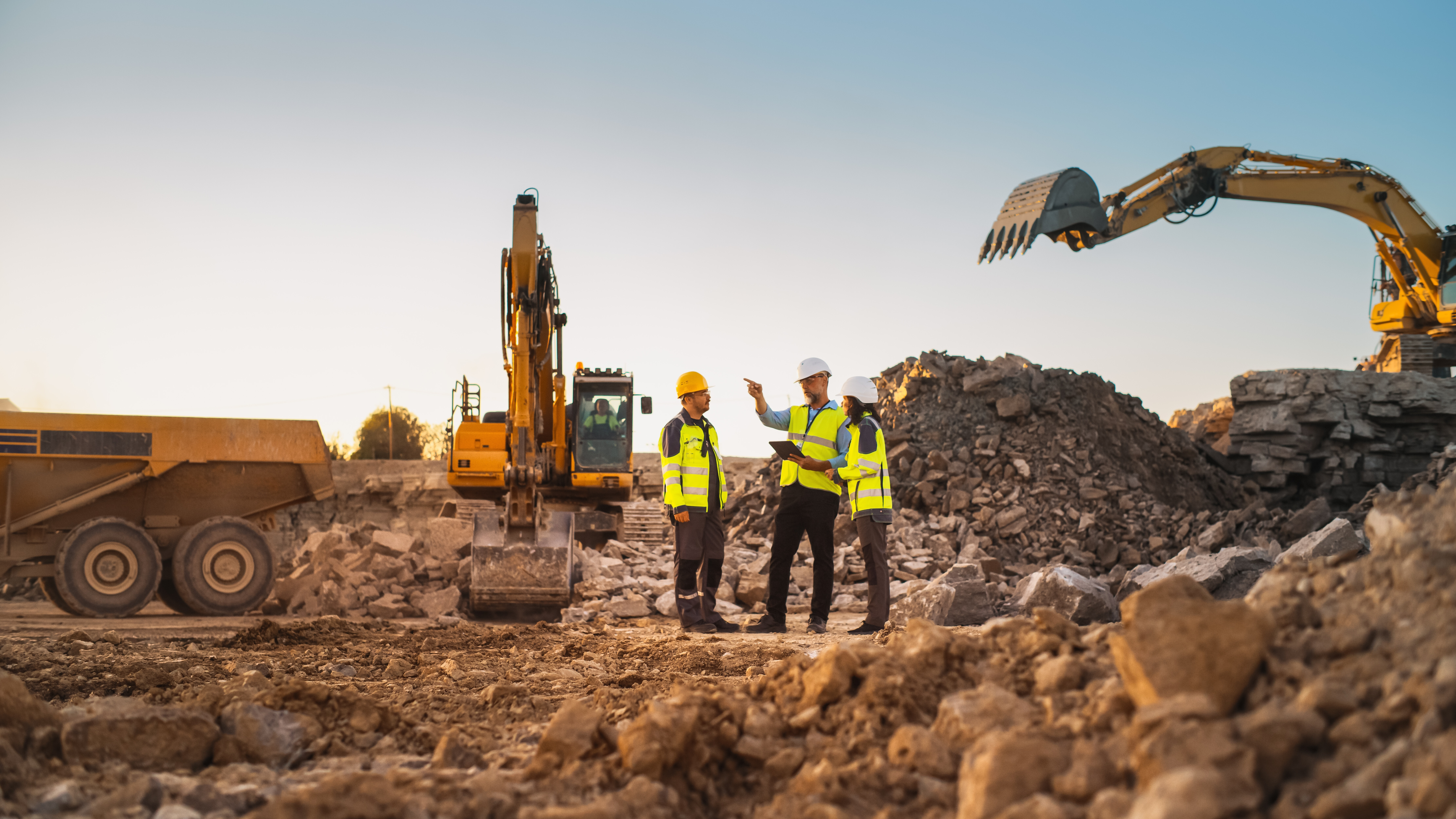 Three workers in safety gear talk at a mining site with excavators and rubble in the background.