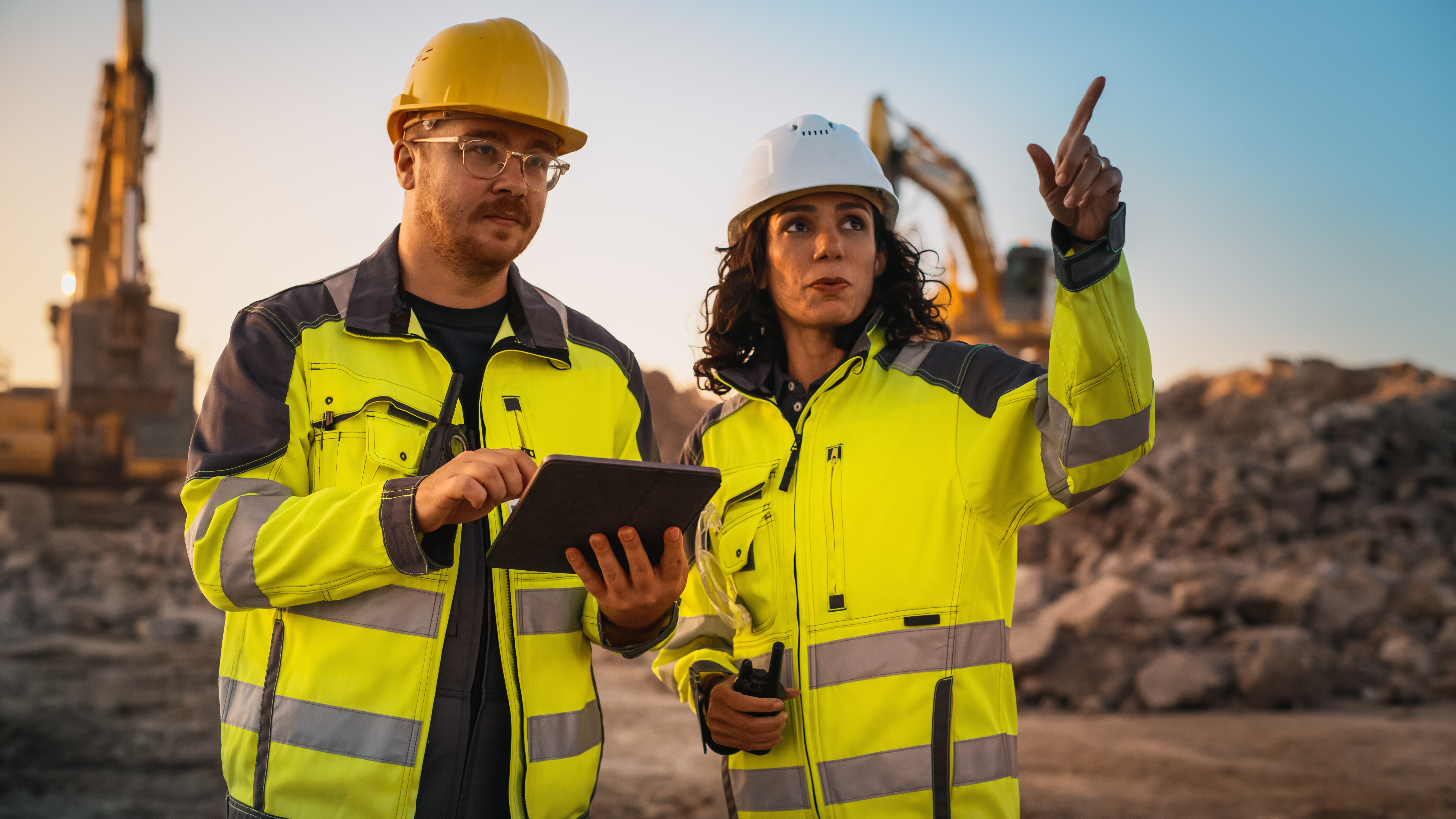 Two workers in safety gear and hard hats discuss at a mining site with excavators in the background.