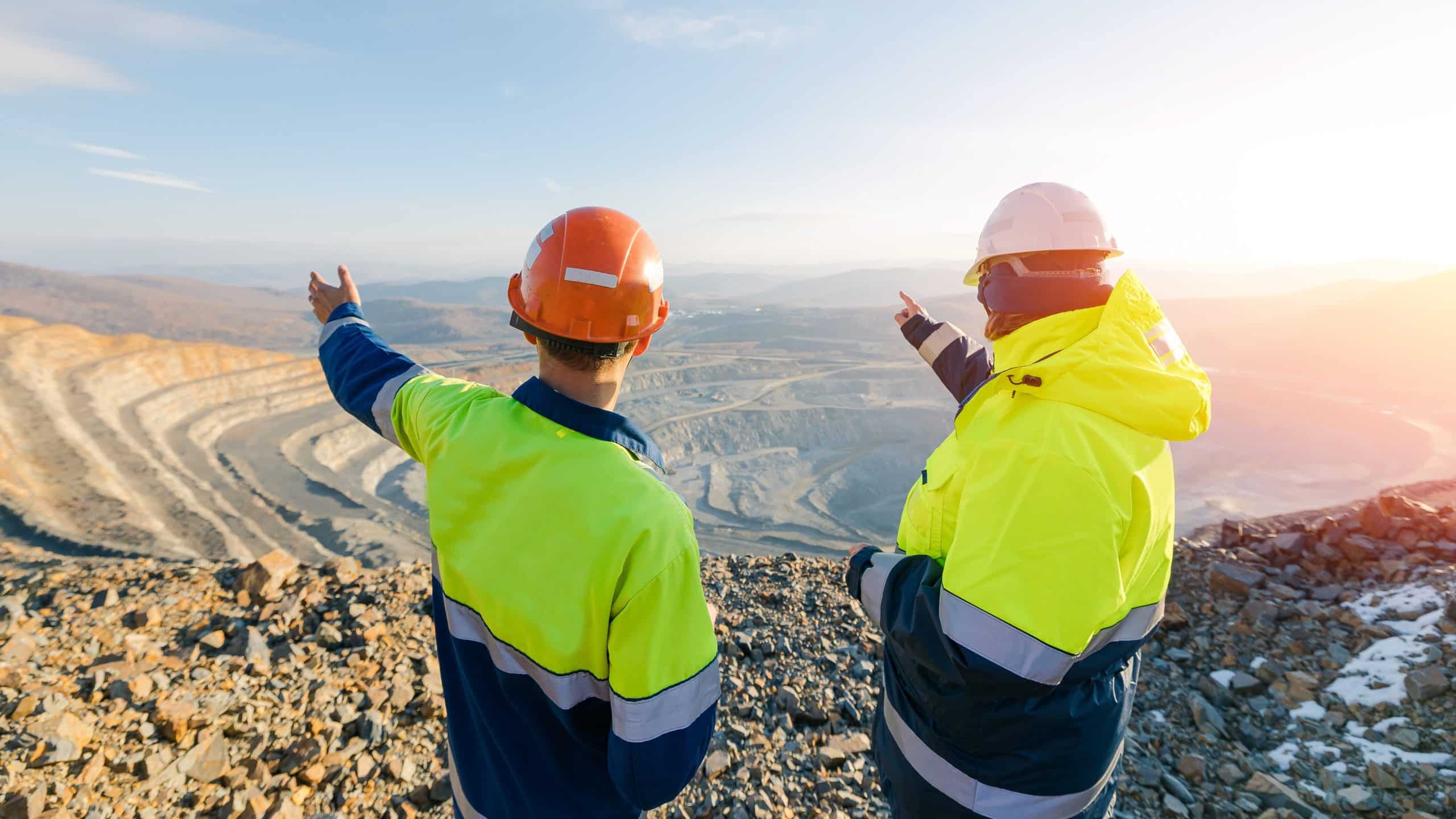 Two workers in safety gear look over and point at a large open-pit mining site in sunlight.