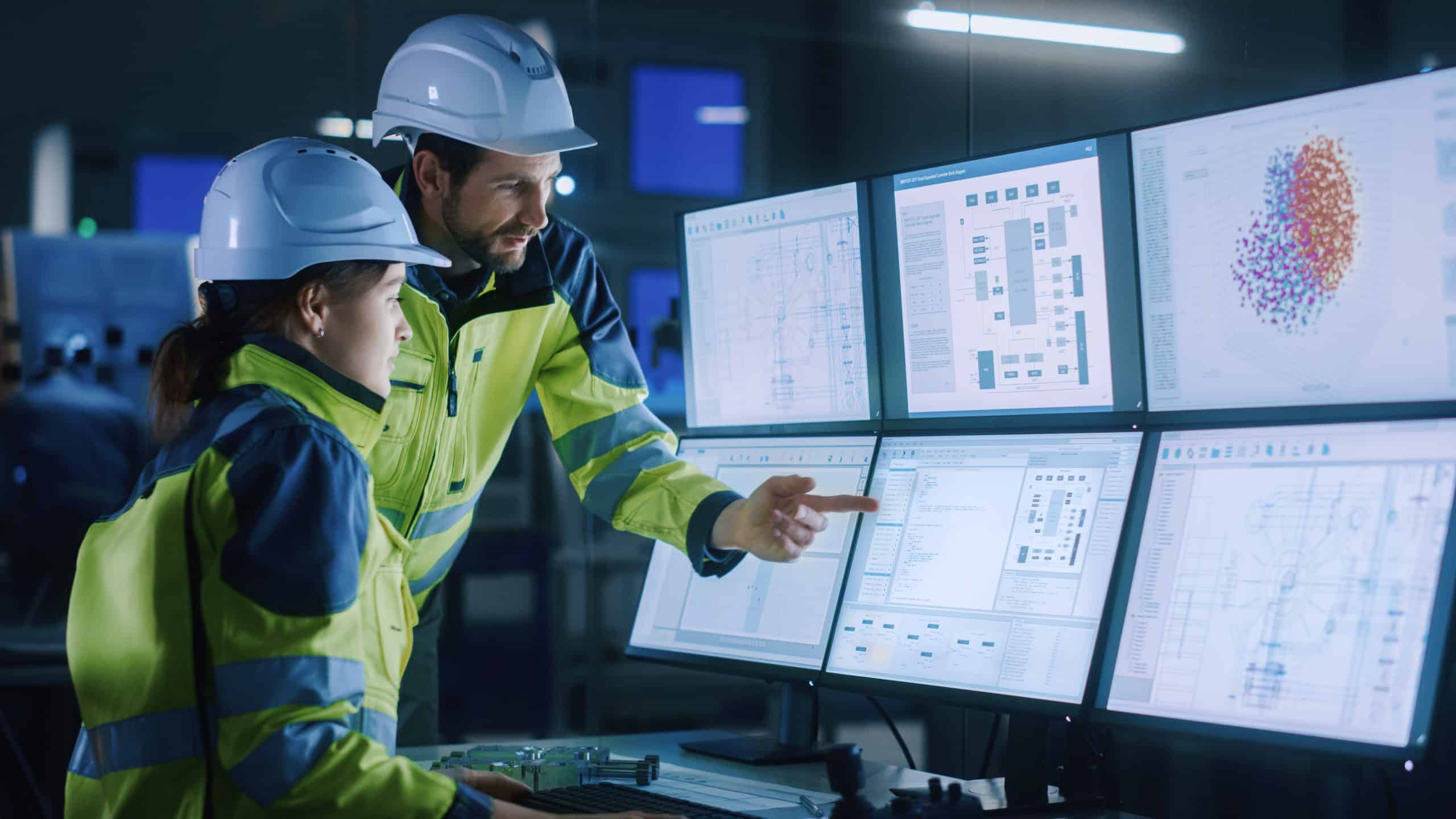 Two engineers in safety gear analyze data on multiple computer monitors in a control room.