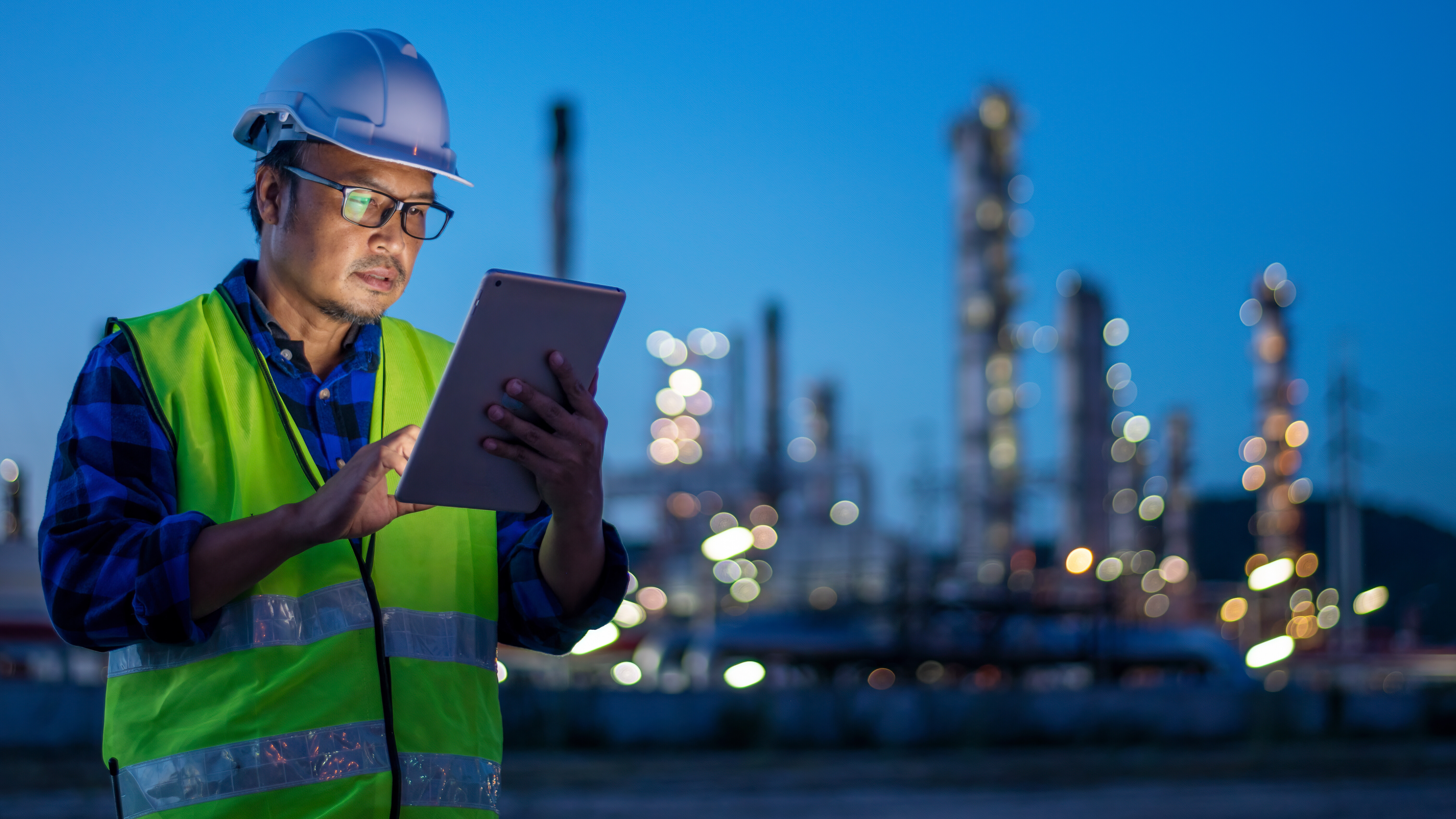 A worker in a hard hat uses a tablet at an industrial site in the evening.