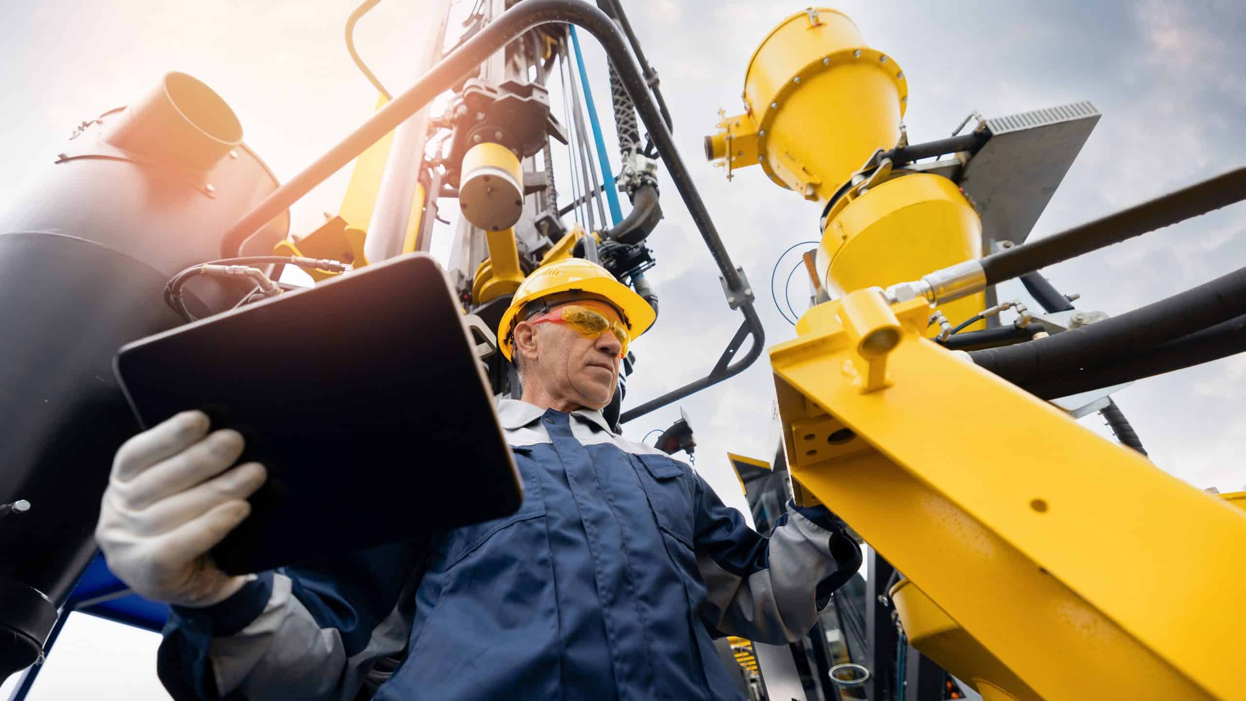 Engineer in safety gear inspects industrial oil and gas equipment while holding a tablet.