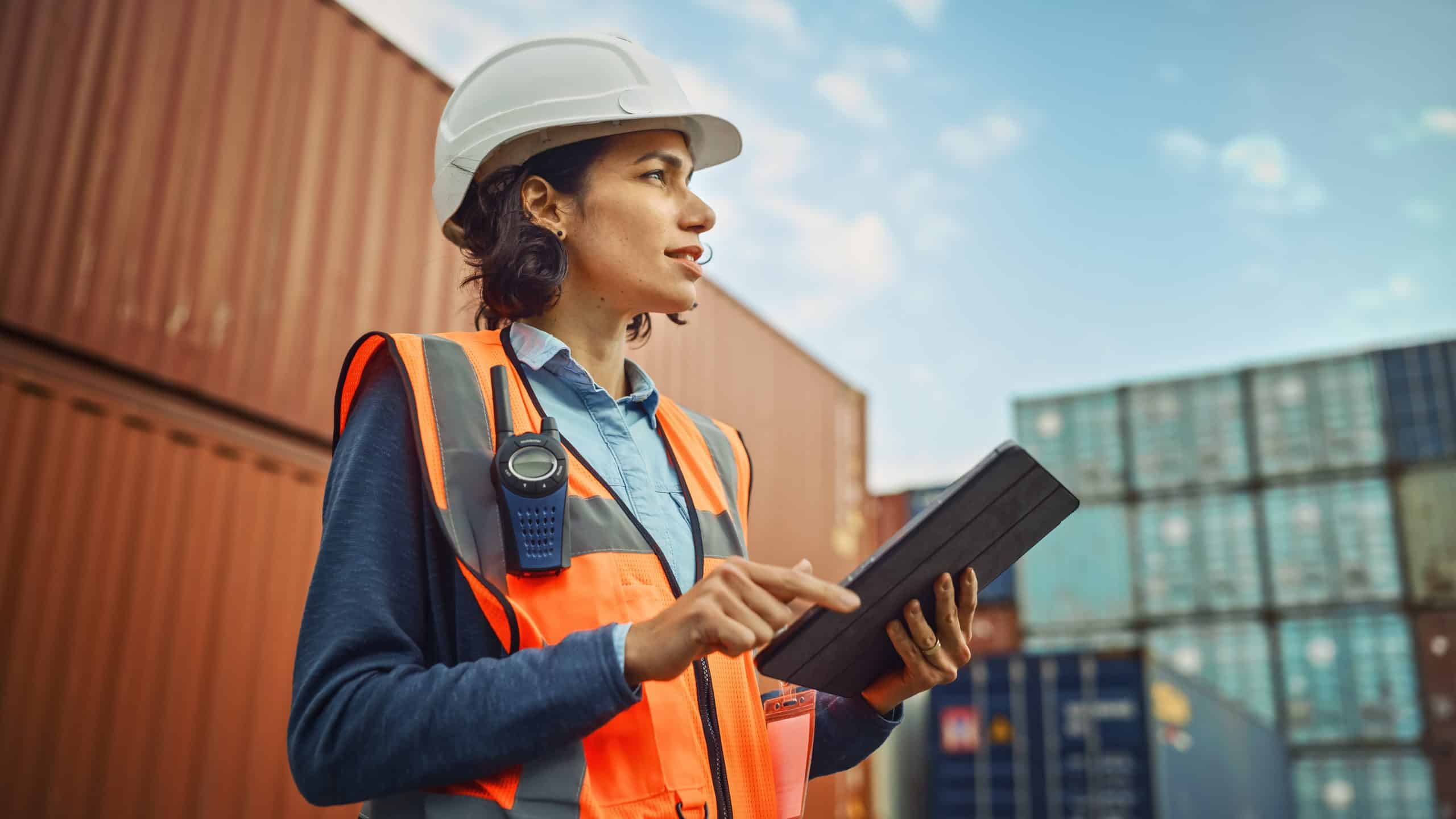 Woman in safety gear using a tablet at a shipping container yard.