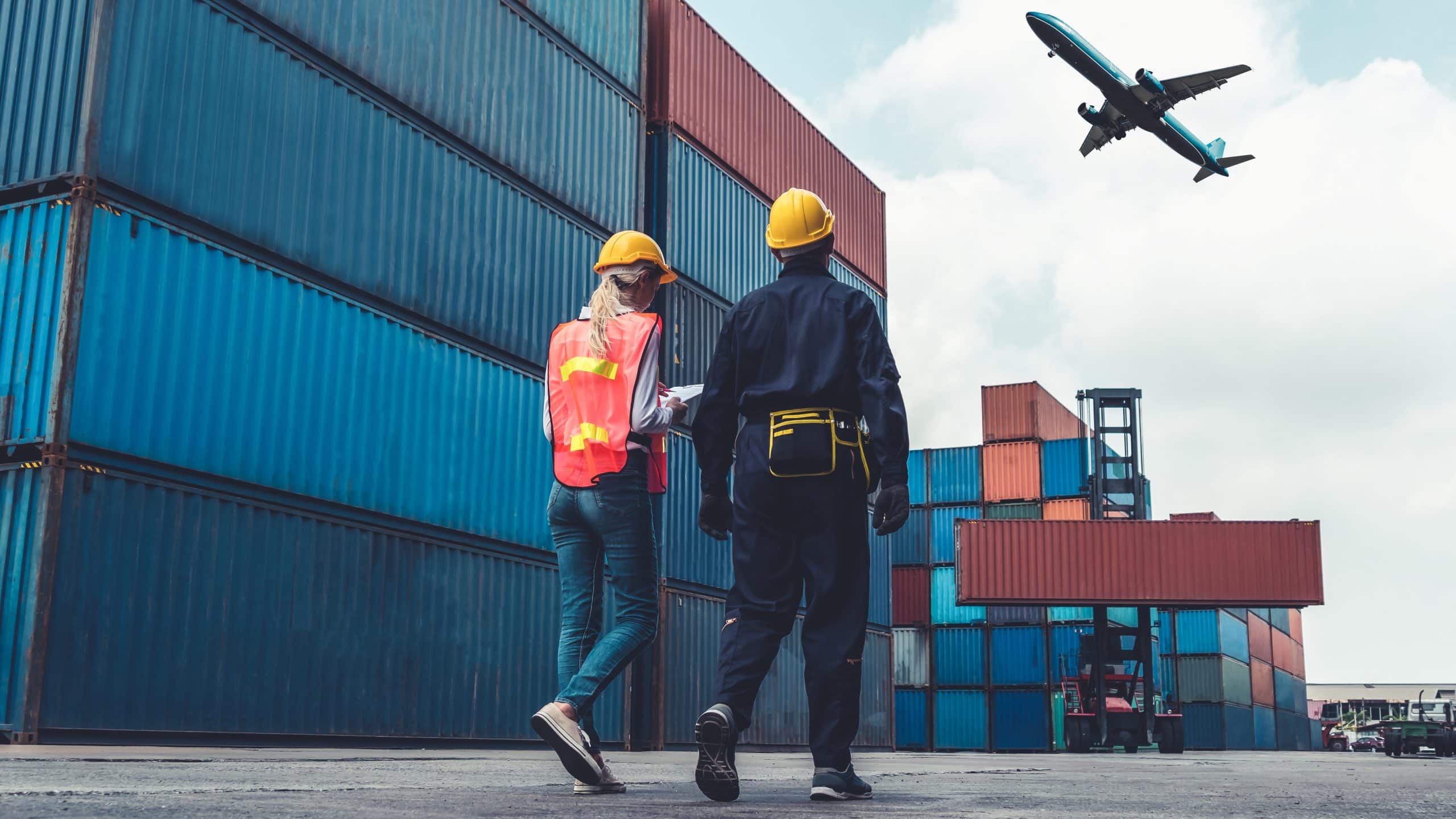Two workers in safety gear at a shipping container yard with an airplane flying overhead.