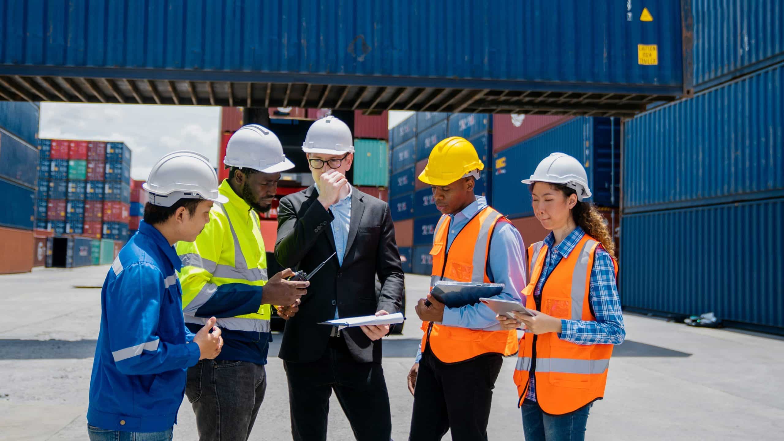 Five workers in safety gear and helmets meet at a shipping container yard.