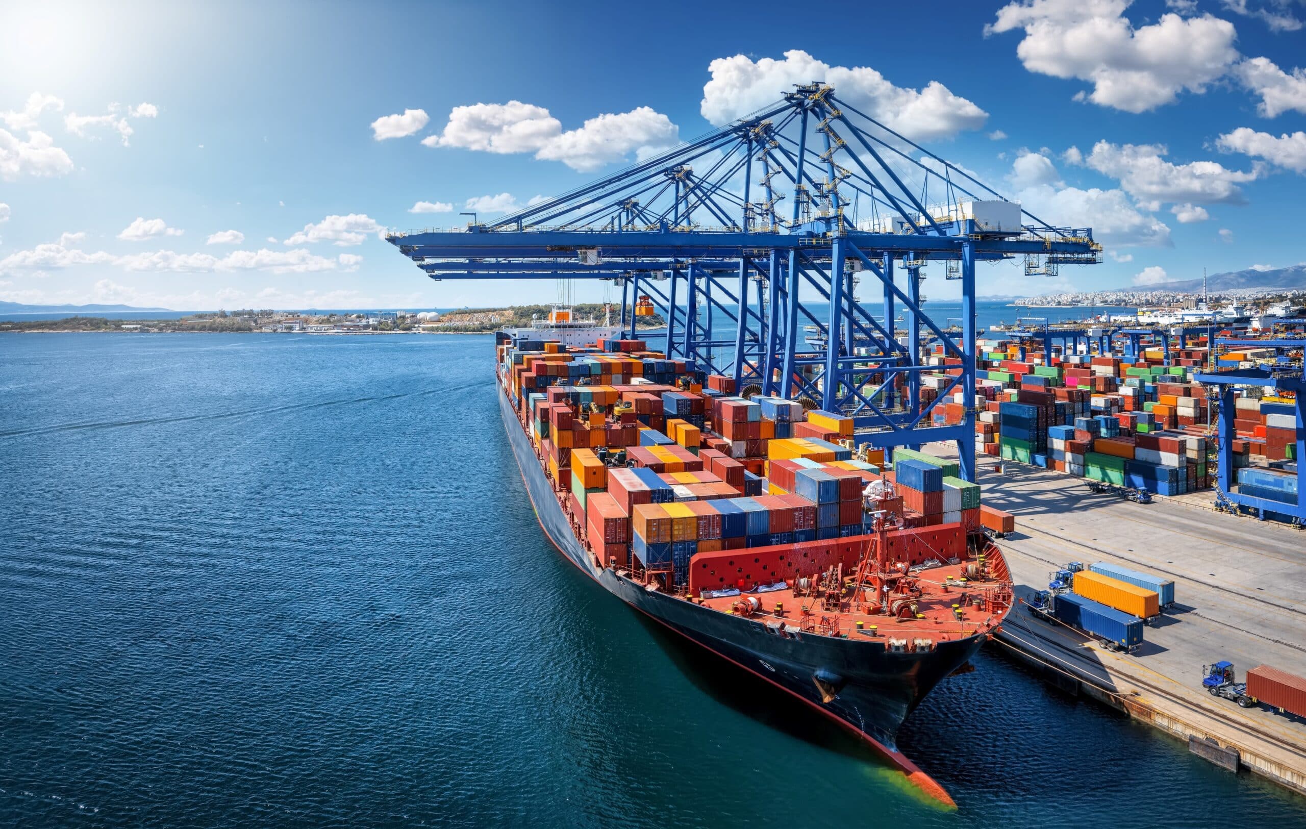 Cargo ship docked at a busy port with shipping containers and large blue cranes.