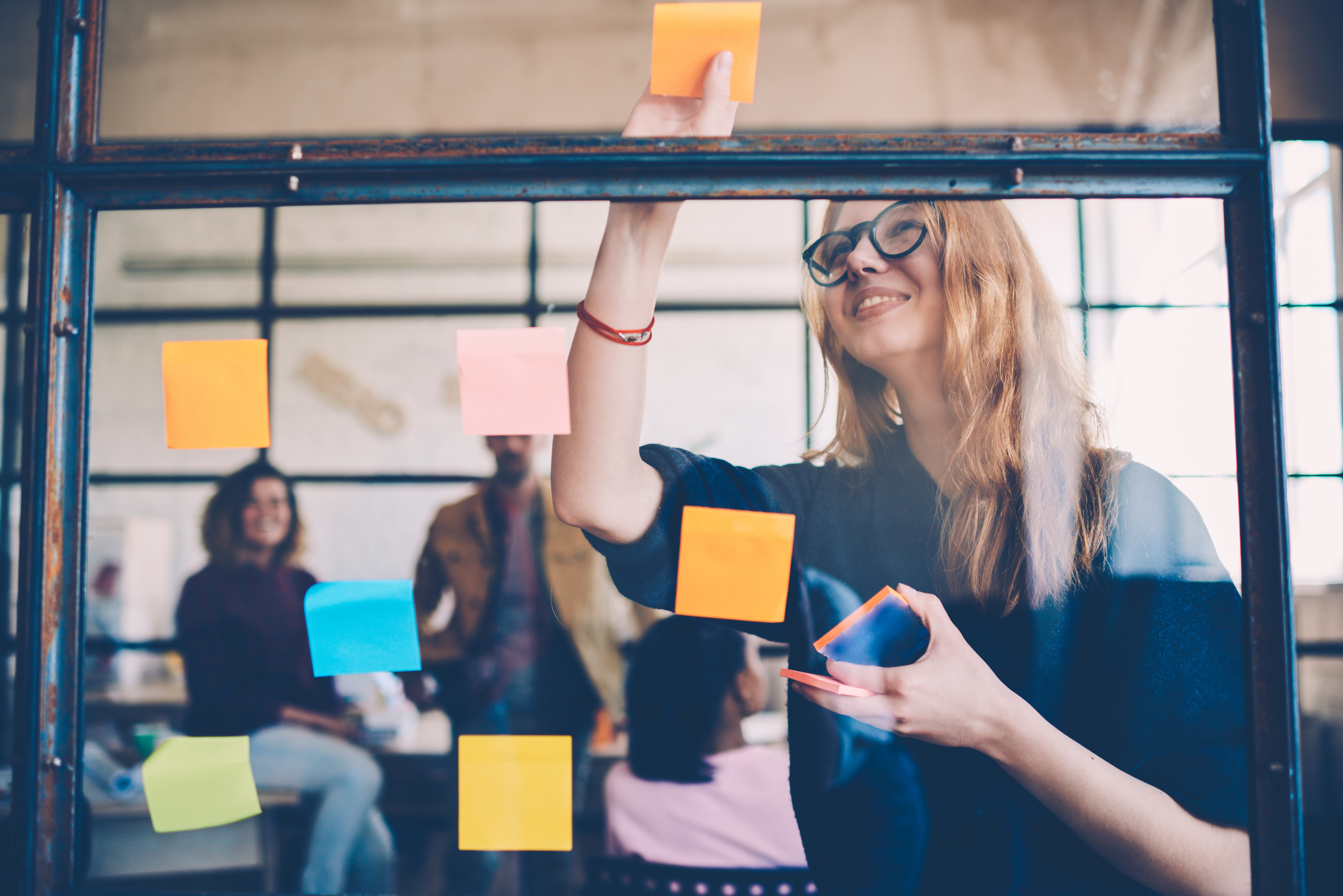 Woman placing sticky notes on glass wall during a meeting with colleagues in the background.