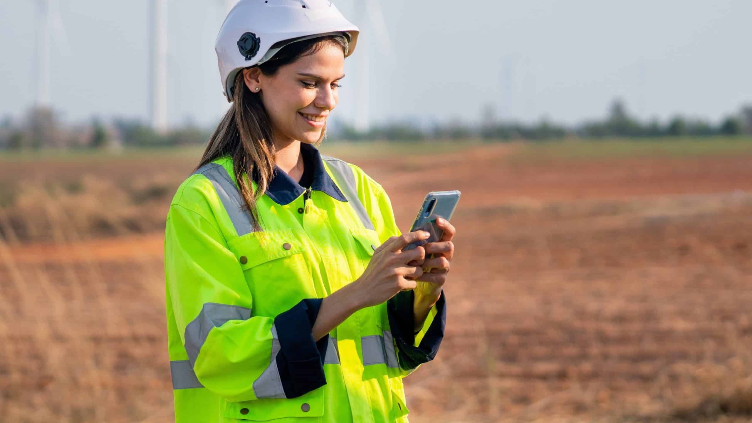 Woman in safety gear and helmet using a smartphone outdoors in a field.