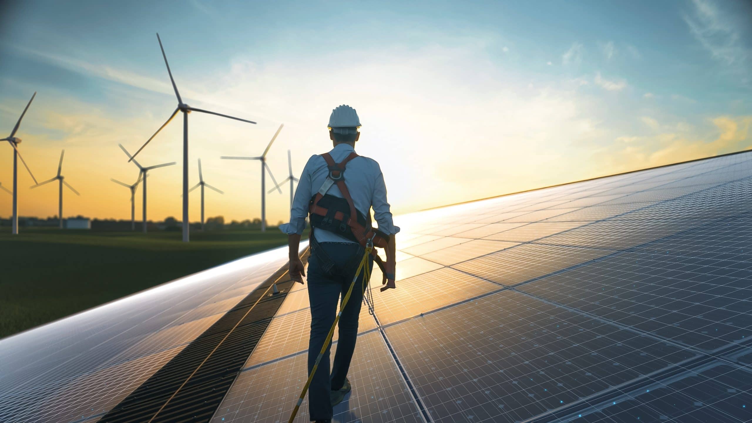 Worker on solar panels at sunset, with wind turbines in the background.