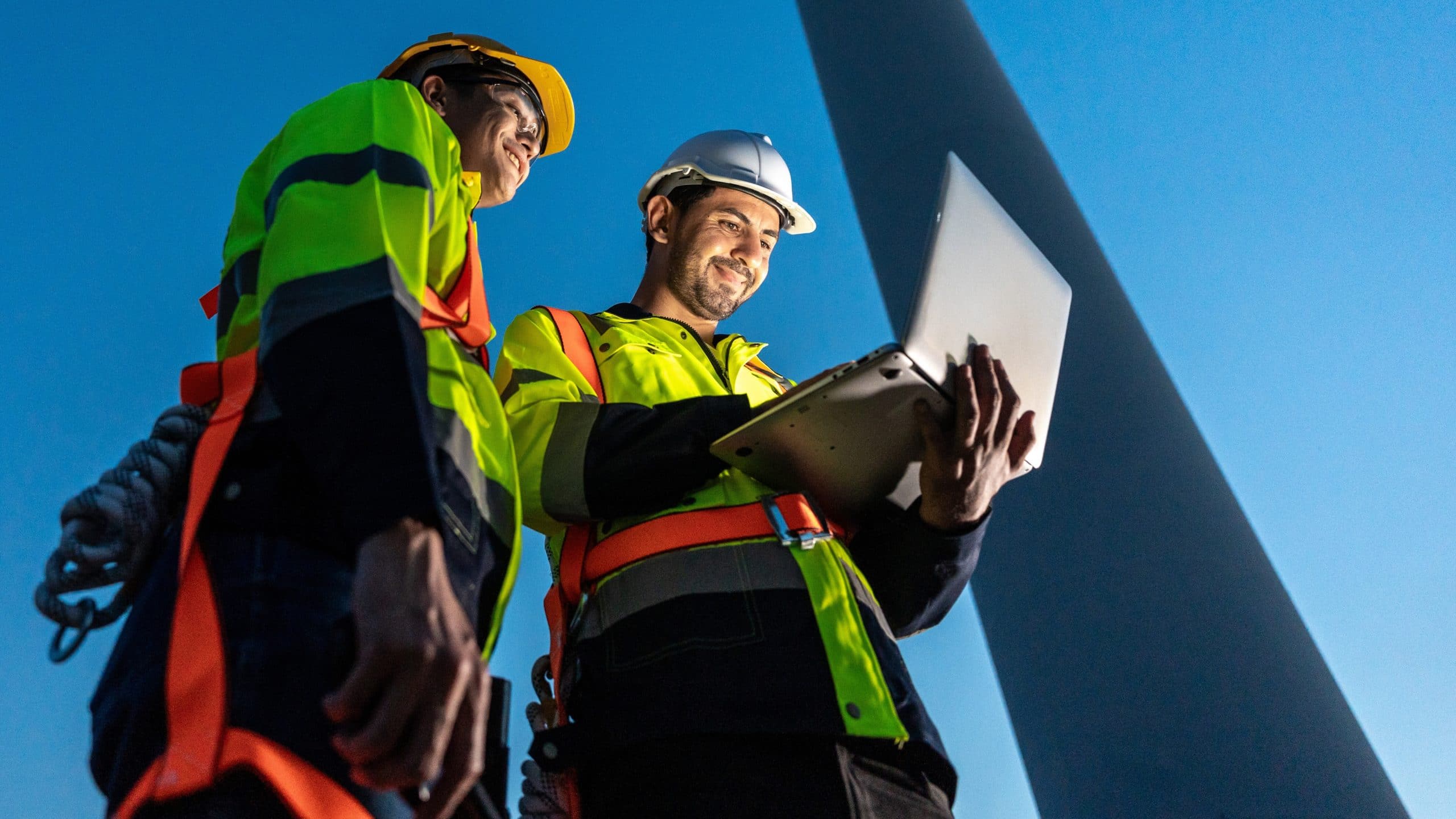 Two engineers in safety gear look at a laptop near a wind turbine at sunset.