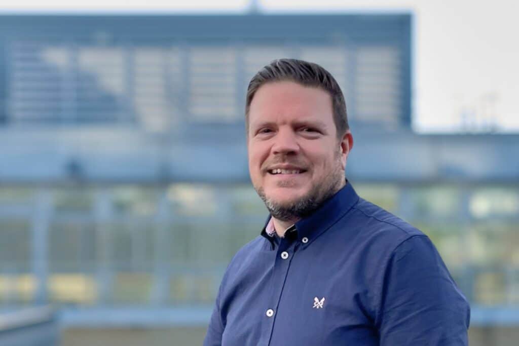 Smiling man in a blue shirt standing outdoors with a modern building in the background.