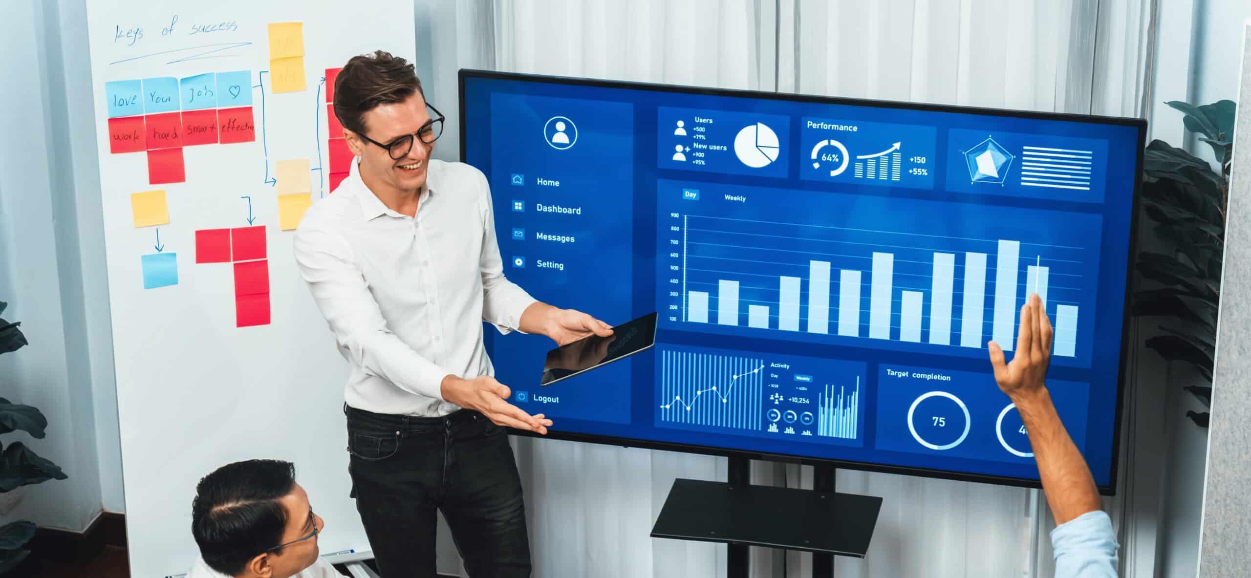 Man presenting data charts on a large screen to colleagues in an office meeting room.