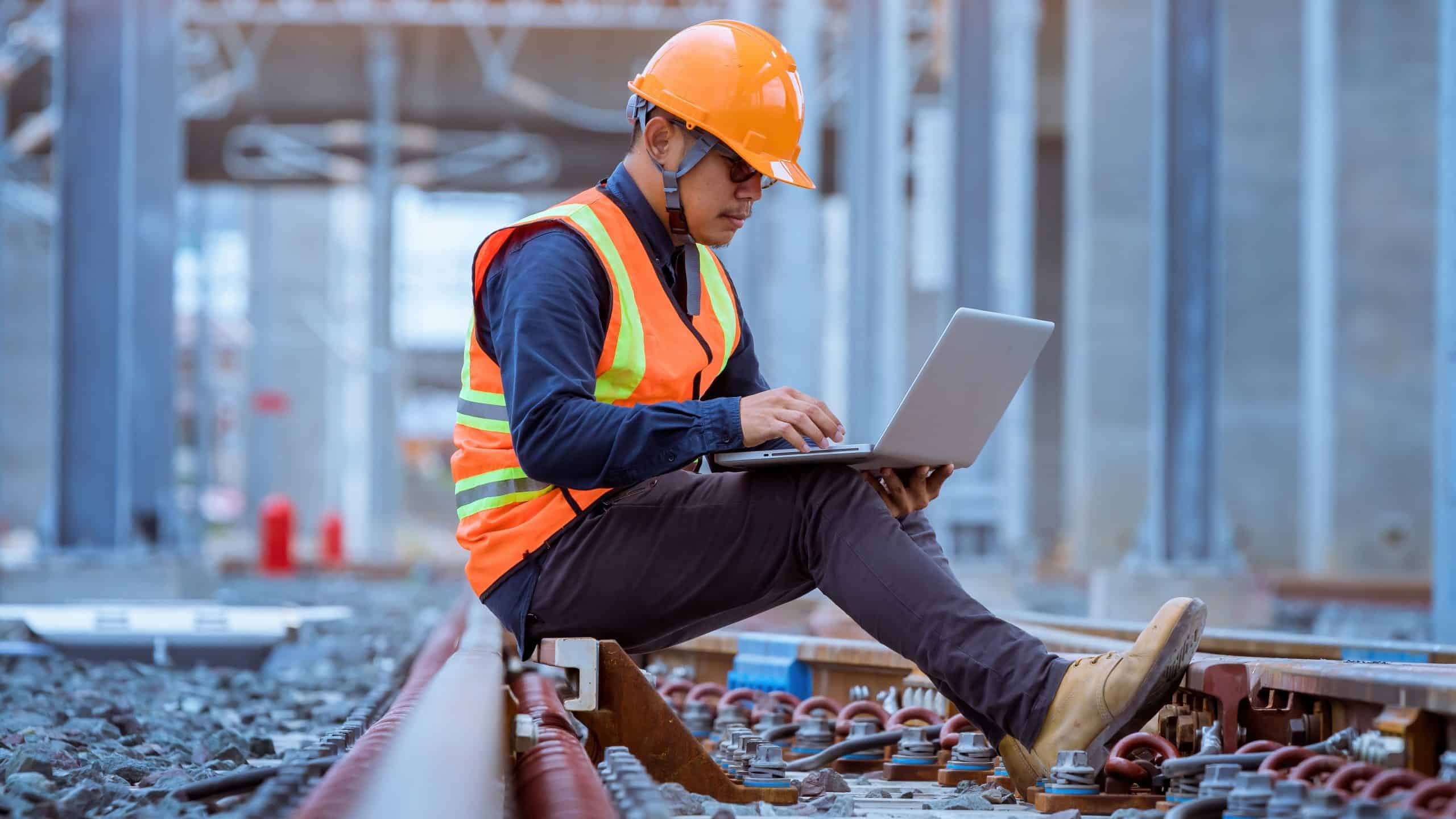 Worker in safety gear sits on train tracks using a laptop at a railway construction site.
