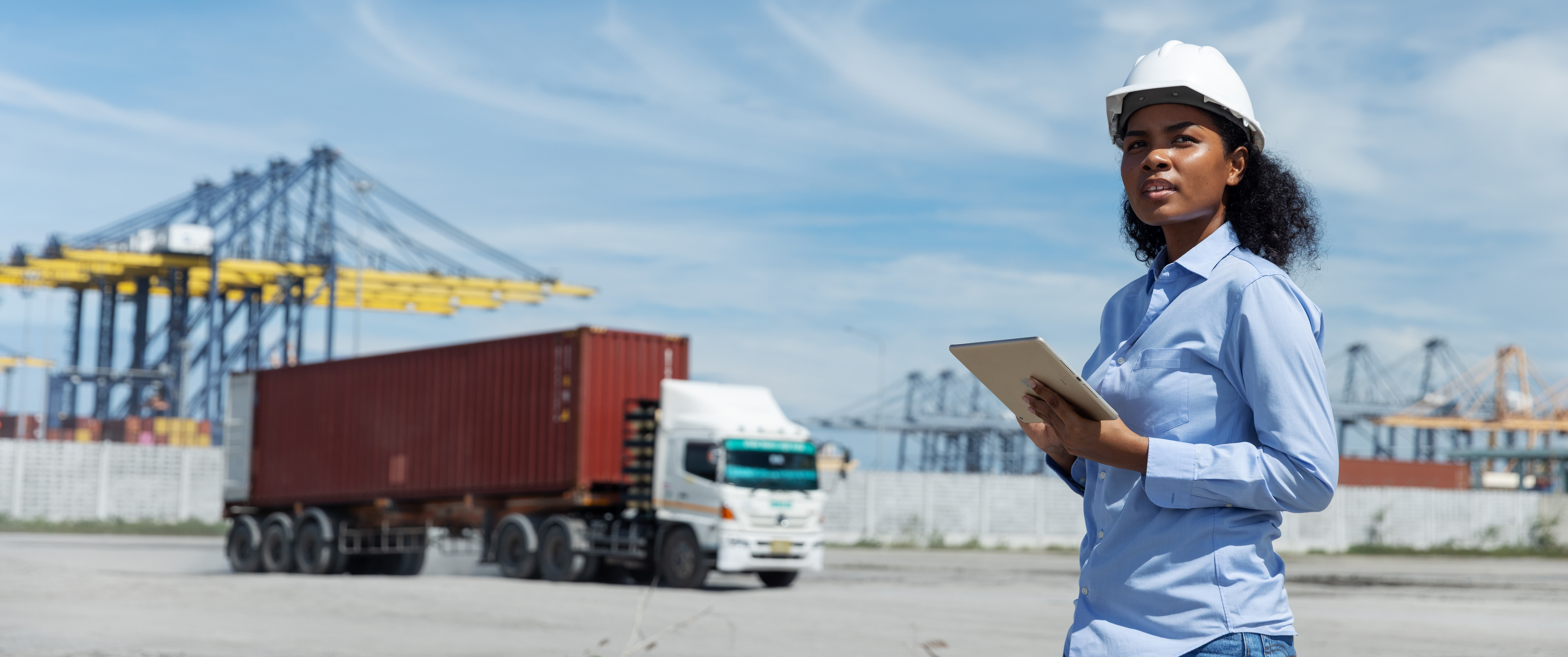 Woman in hard hat with tablet at shipping port, truck and containers in background.
