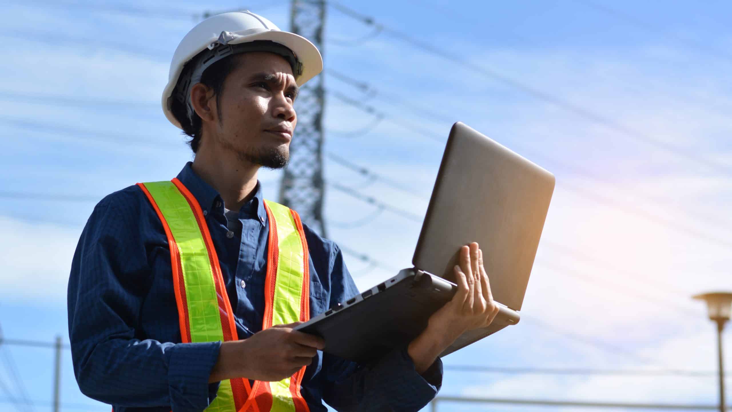 Engineer with a hard hat and safety vest using a laptop outdoors near power lines.