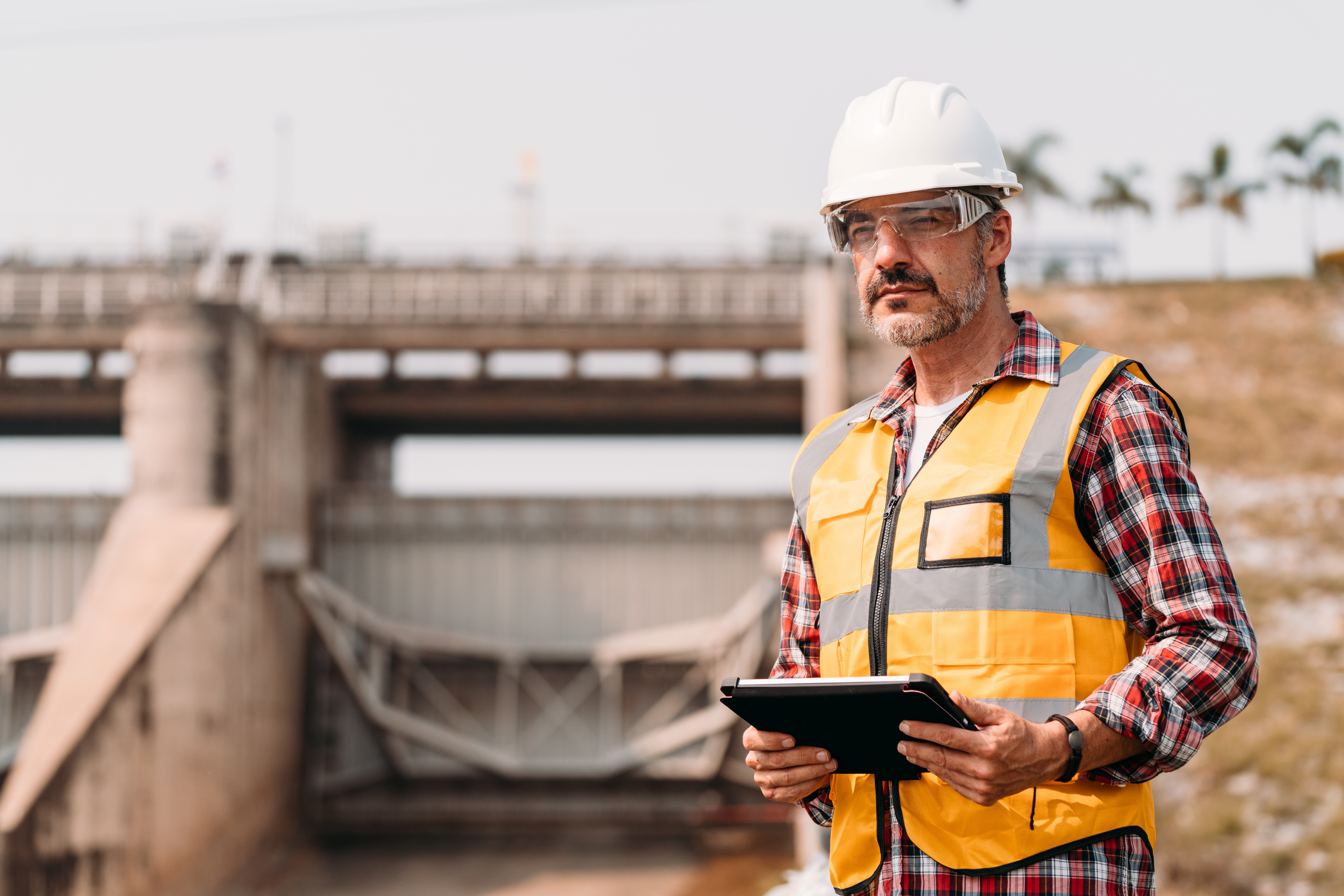 Engineer in safety gear holds tablet at a power plant facility.