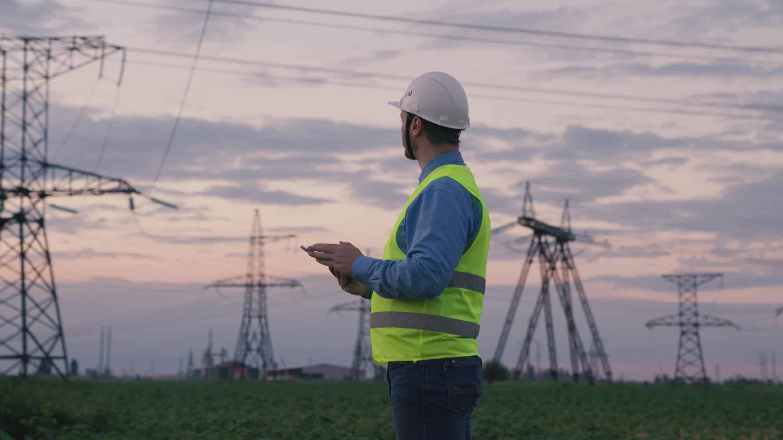 A worker in a hard hat and vest surveys power lines at sunset.