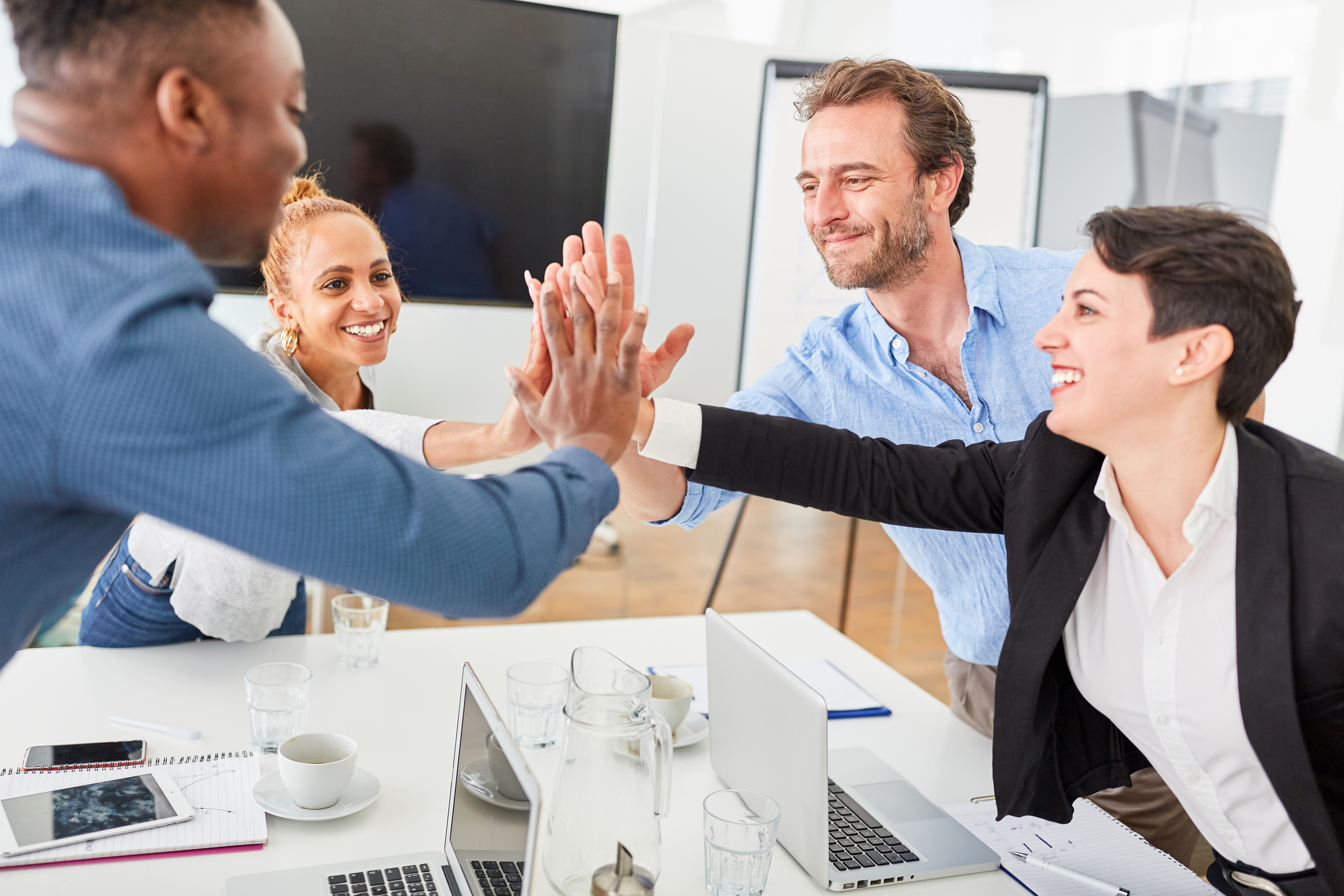 Four coworkers smiling and giving a group high five in an office meeting.