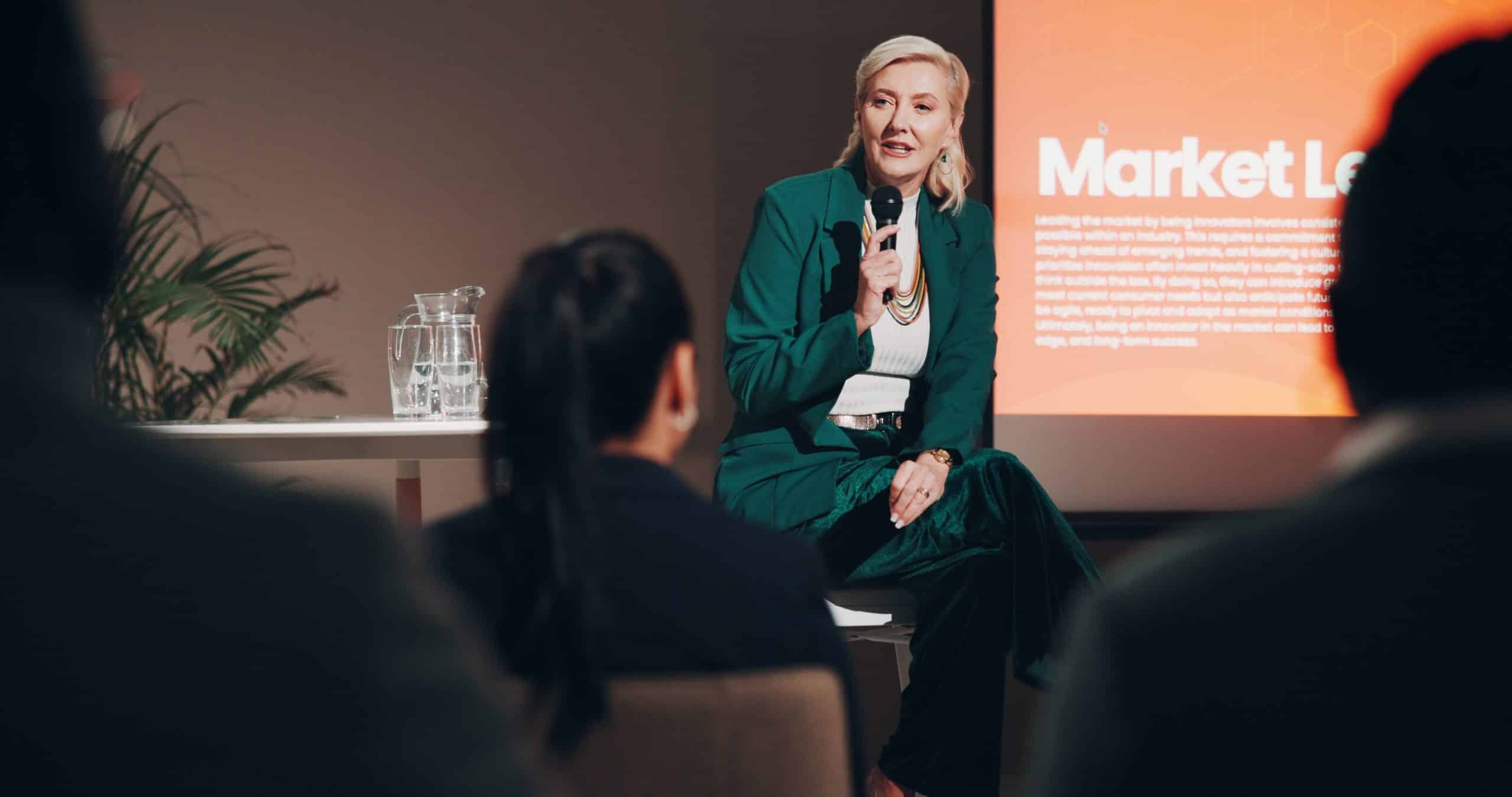A woman speaks into a microphone during a presentation with an audience and screen in the background.