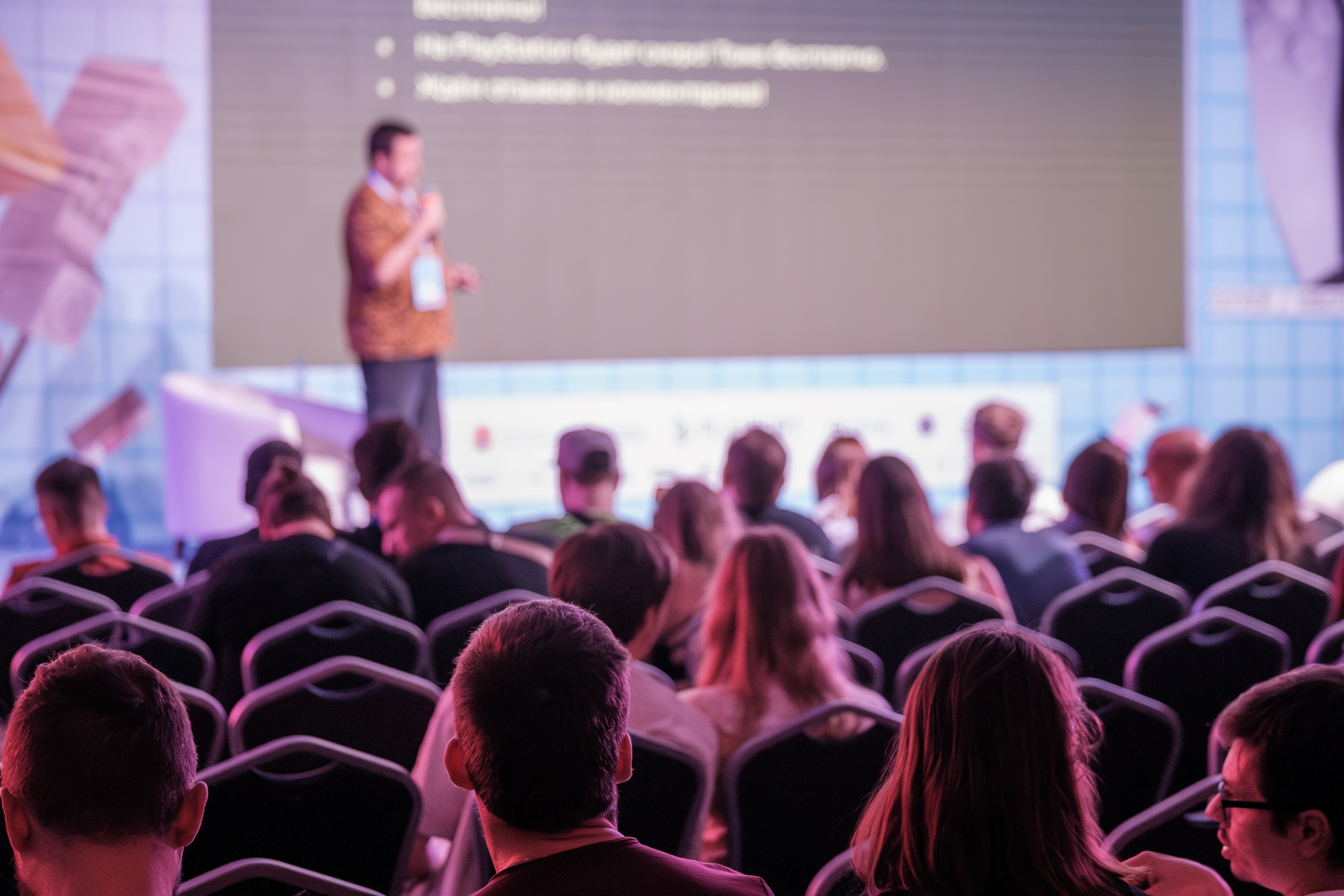 People sitting in an audience watching a speaker give a presentation on stage.