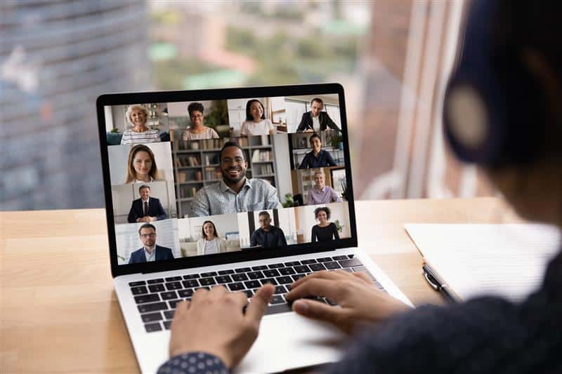 Person on laptop in a video conference call with multiple participants visible on the screen.