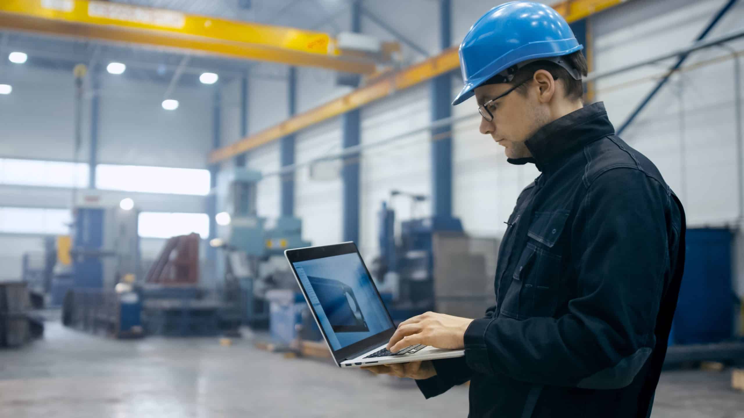 Man in a blue hard hat using a laptop in a large industrial warehouse.