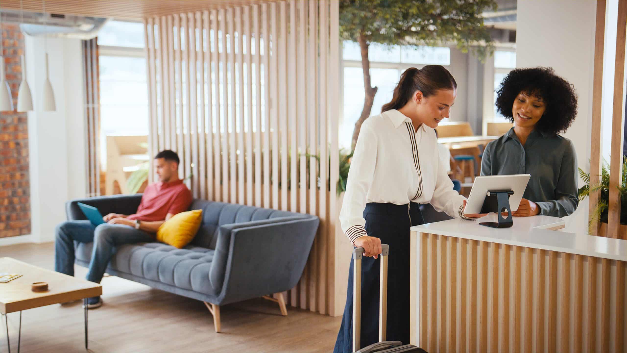 Woman checks in at a front desk; another woman assists while a man works on a laptop in the background.