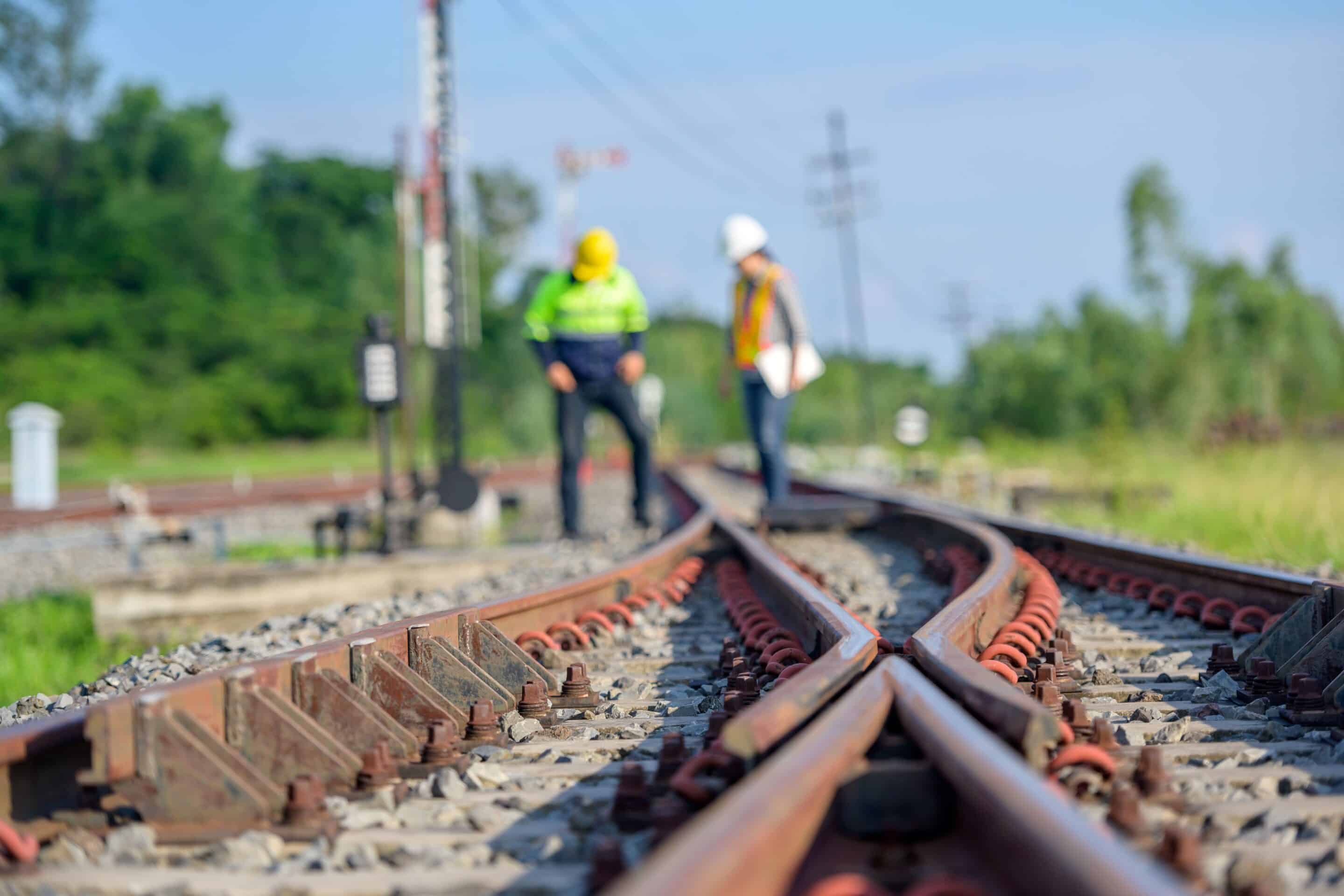 Two workers in safety gear inspect railway tracks outdoors.